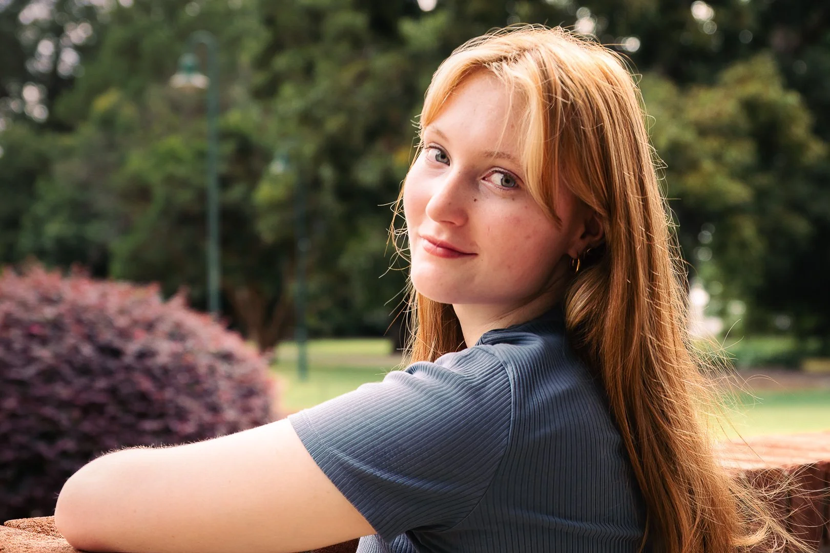 A young woman with long red hair and blue eyes sitting outdoors in a park, looking at the camera with a slight smile.