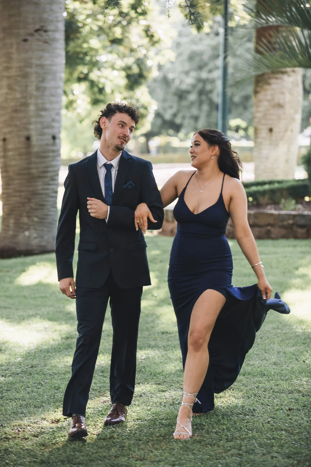 A man and woman dressed in formal attire walking on a grassy area with large trees in the background. The man is wearing a dark suit and tie, and the woman is in a dark blue gown with a high slit looking very natural for their photography session