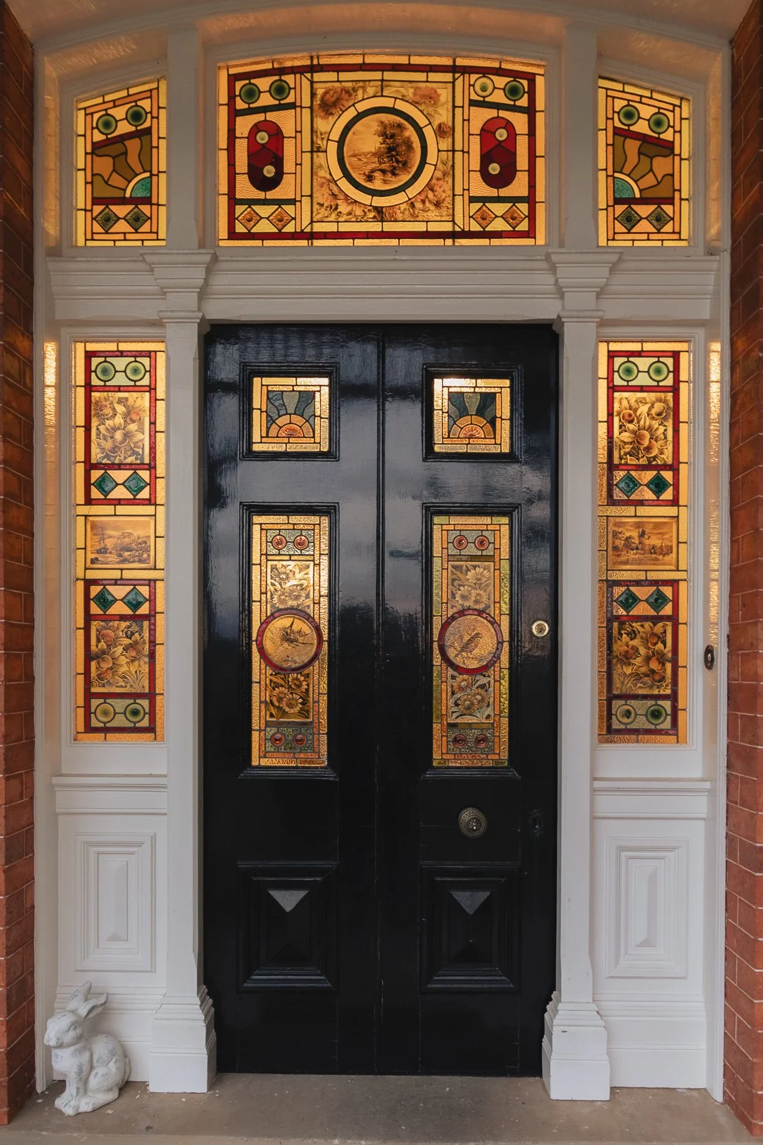 Decorative black front door with stained glass panels featuring floral and landscape designs, framed by white trim and side panels with similar stained glass, and brick walls on either side. Captured by accommodation photographer, Toowoomba