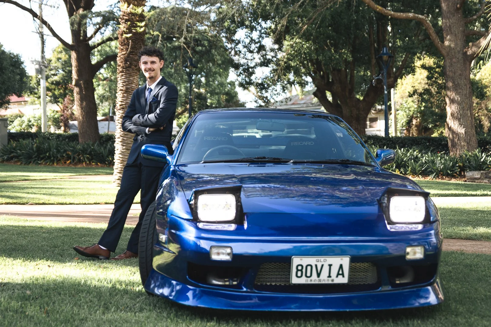 A man in a dark suit stands with arms crossed next to a blue sports car on a grassy yard, with trees and houses in the background.