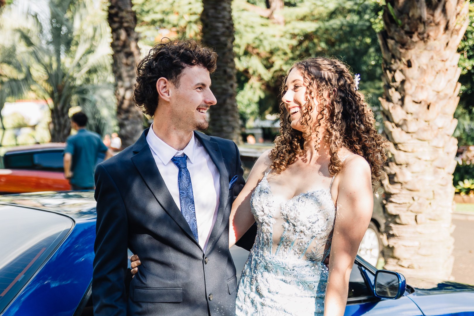 A smiling man in a dark suit and a woman in a wedding dress share a happy moment outdoors in front of palm trees.