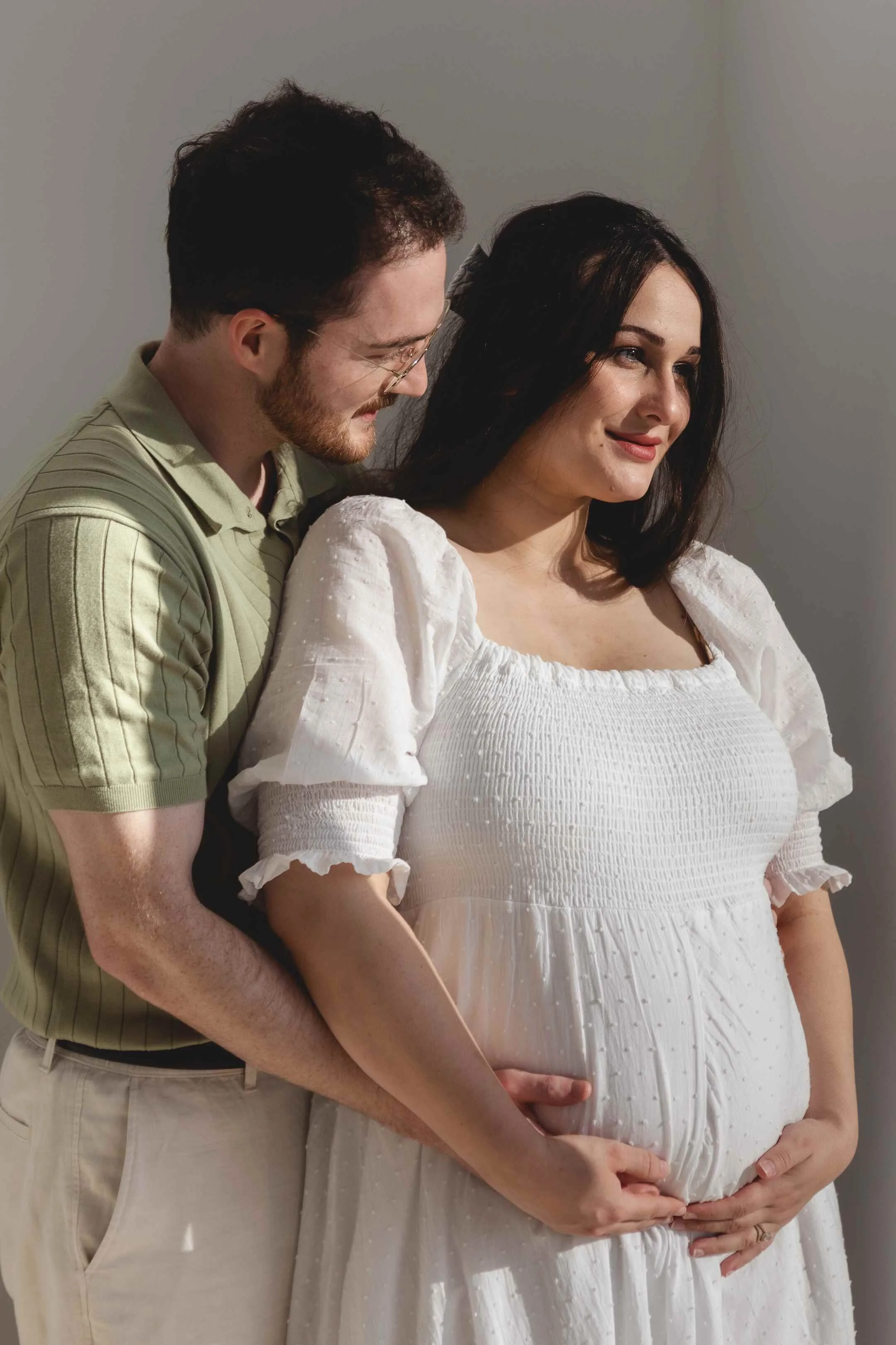 A couple with light skin, the man with glasses and a beard, cuddles with a pregnant woman in a white dress against a neutral background.