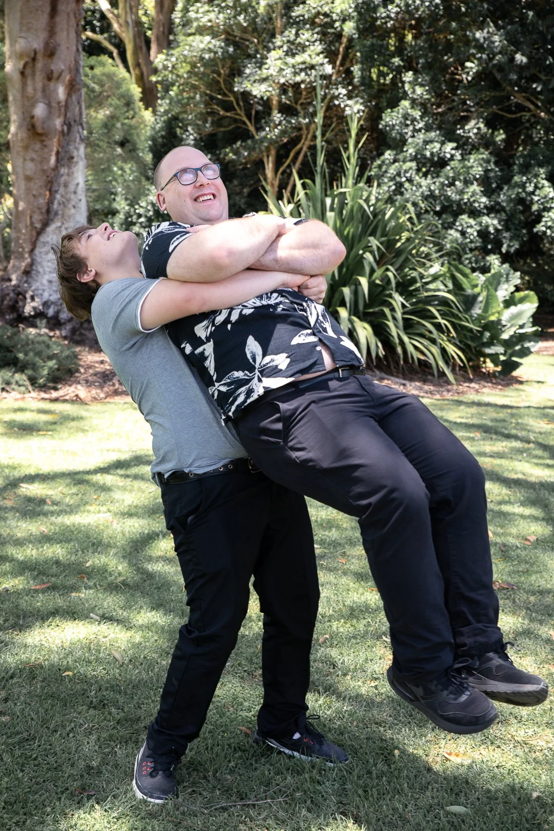 Two men outdoors, one lifting the other in a playful embrace, smiling and laughing, with trees and greenery in the background.
