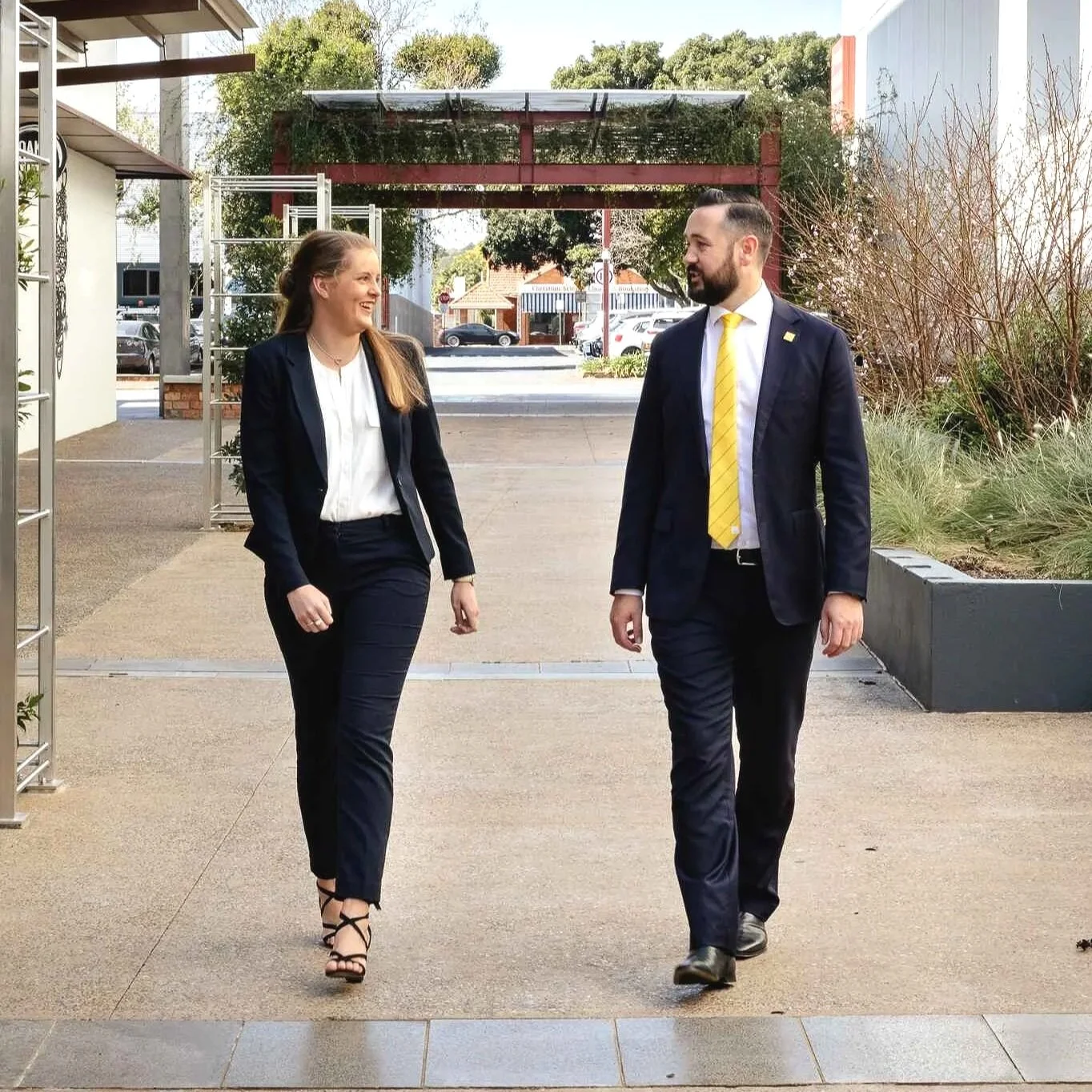 Two professionally dressed people, a woman and a man, walking outdoors and smiling at each other.