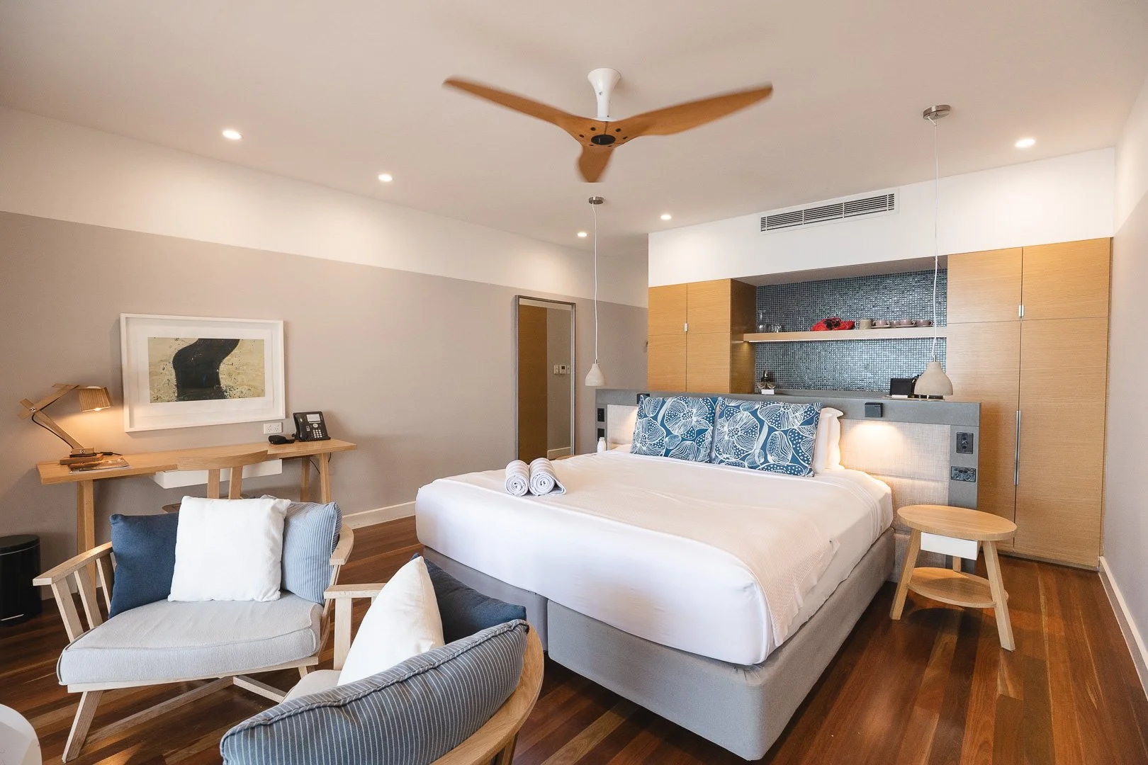 Modern bedroom with a large bed, blue patterned pillows, wooden furniture, a small seating area, a desk with a lamp, and a ceiling fan. Beach Club Hotel, Hamilton Island Whitsundays. Photography by Laurene Bardsley Photography