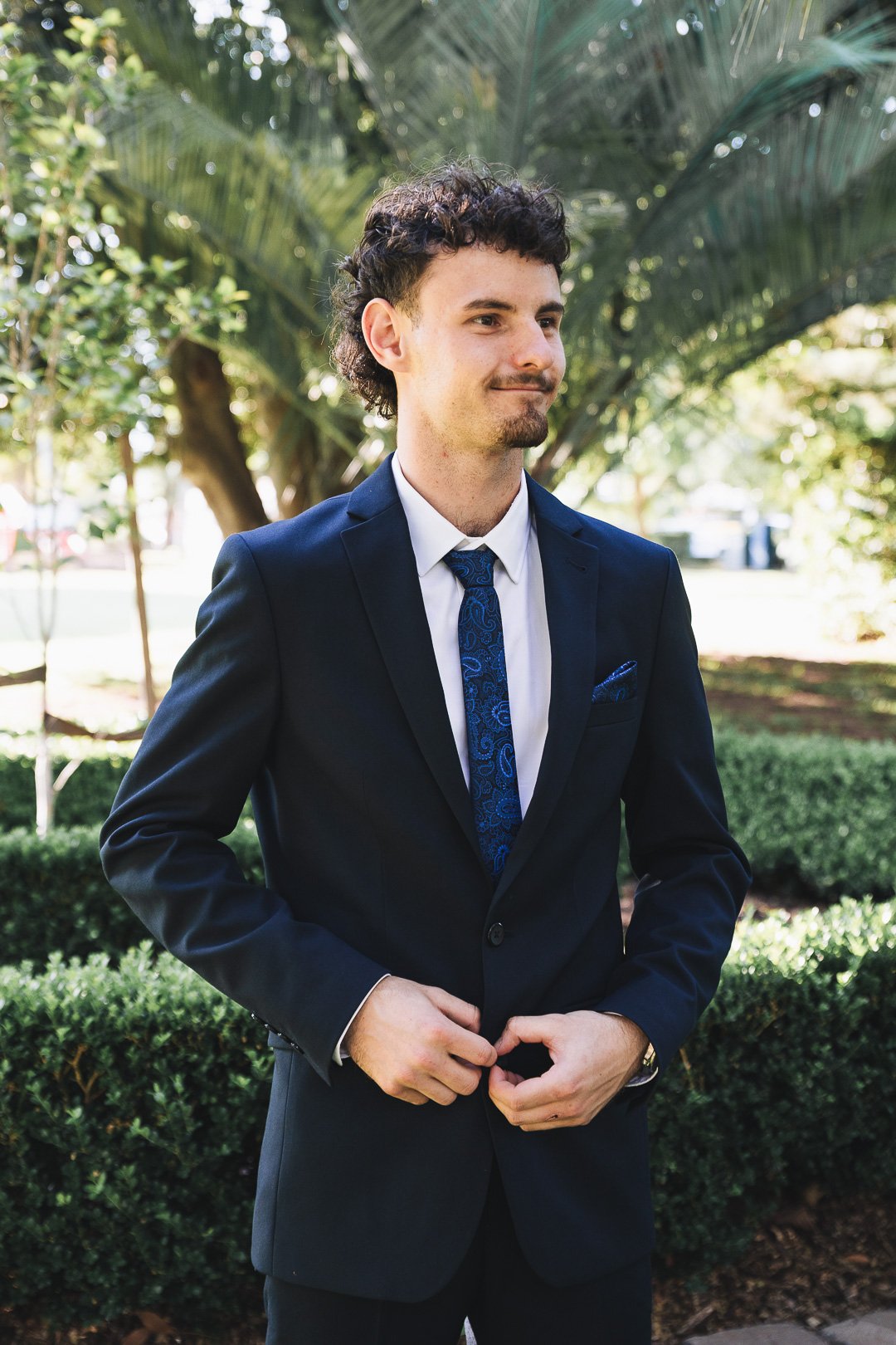 A young man in a navy suit, white shirt, and blue patterned tie stands outdoors among trees and bushes, adjusting his suit jacket.