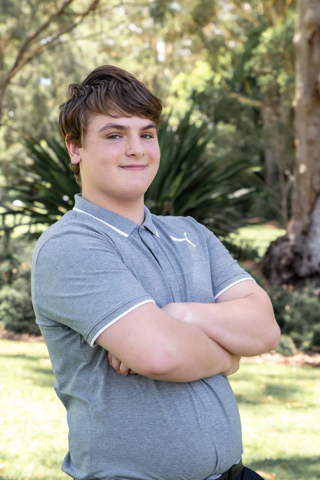 A young boy with brown hair, wearing a gray polo shirt, standing outdoors with arms crossed, in front of green trees and bushes.