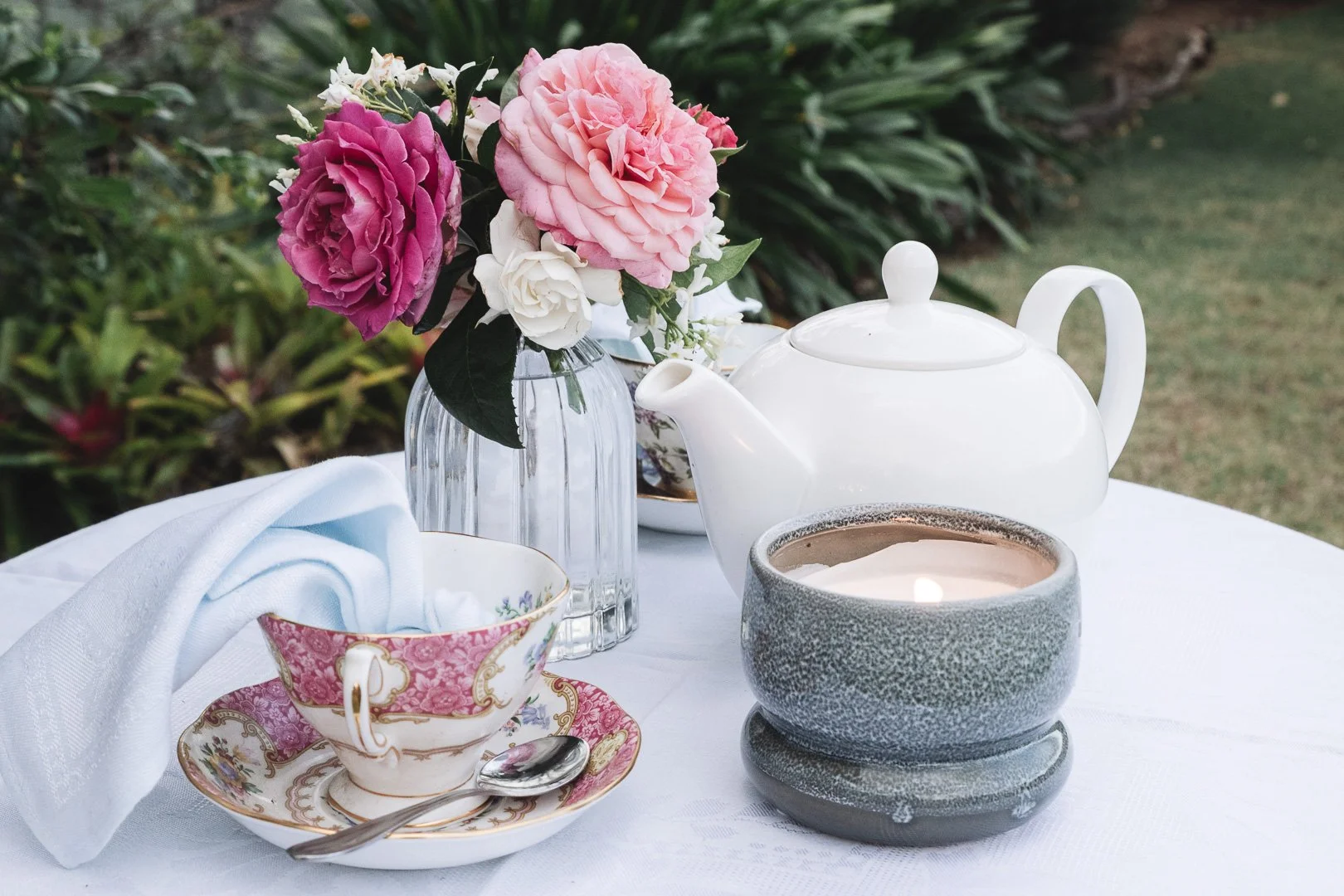 A teapot, a cup with a saucer and a spoon, a lit candle in a textured gray holder, and a vase of pink and white flowers are arranged on a white tablecloth outdoors.