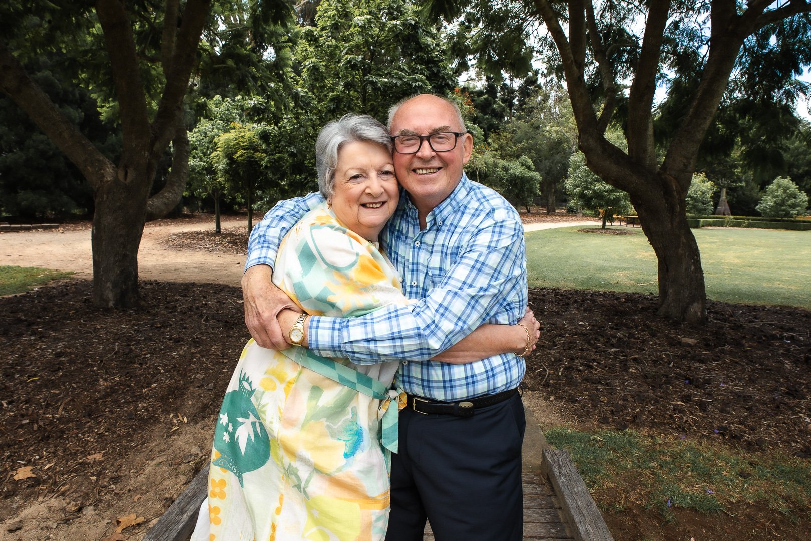 A happy elderly woman and man embracing each other outdoors in a park with trees and grass, smiling at the camera.