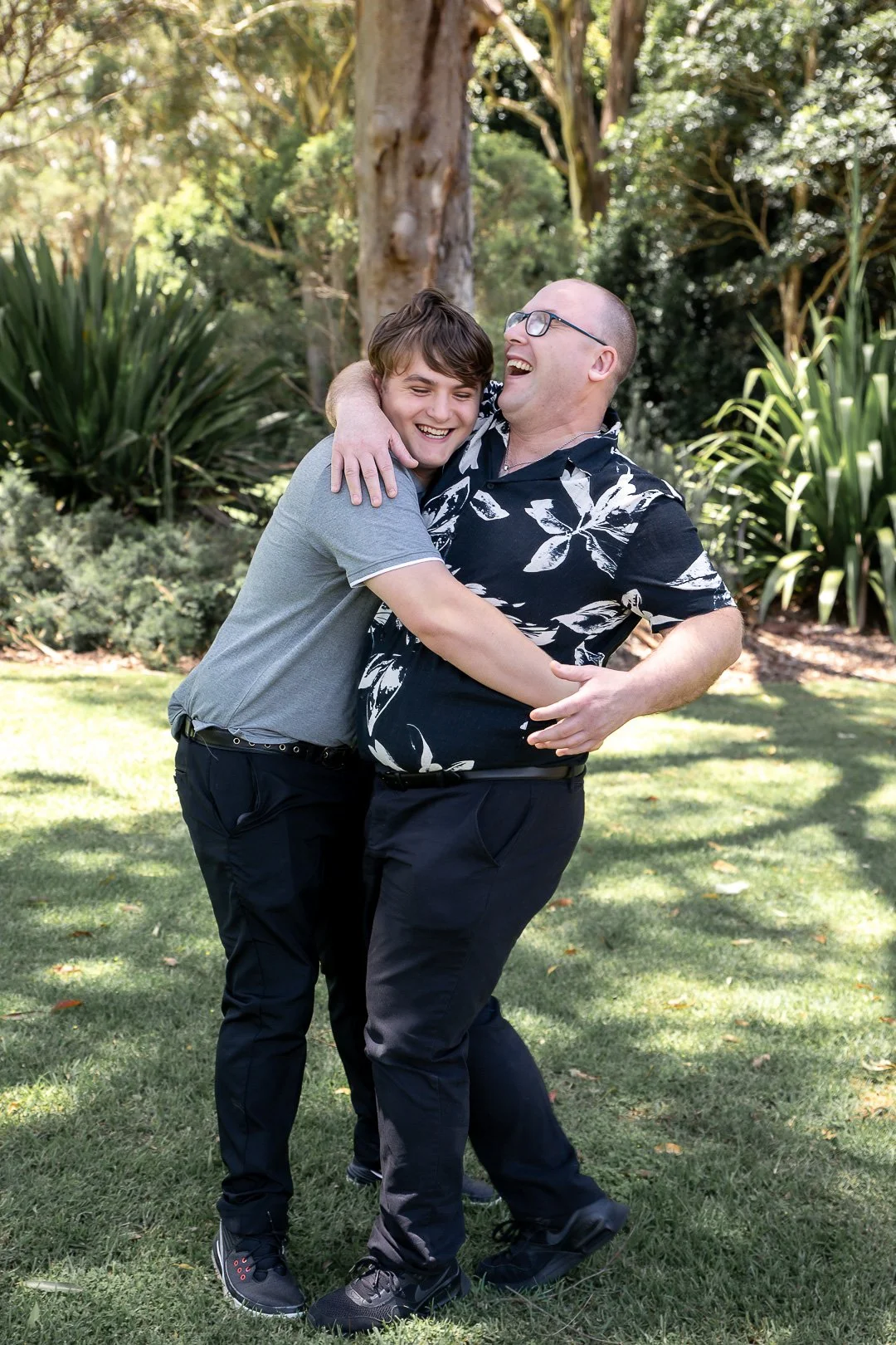 Two men are embracing and laughing in a park surrounded by trees and greenery.