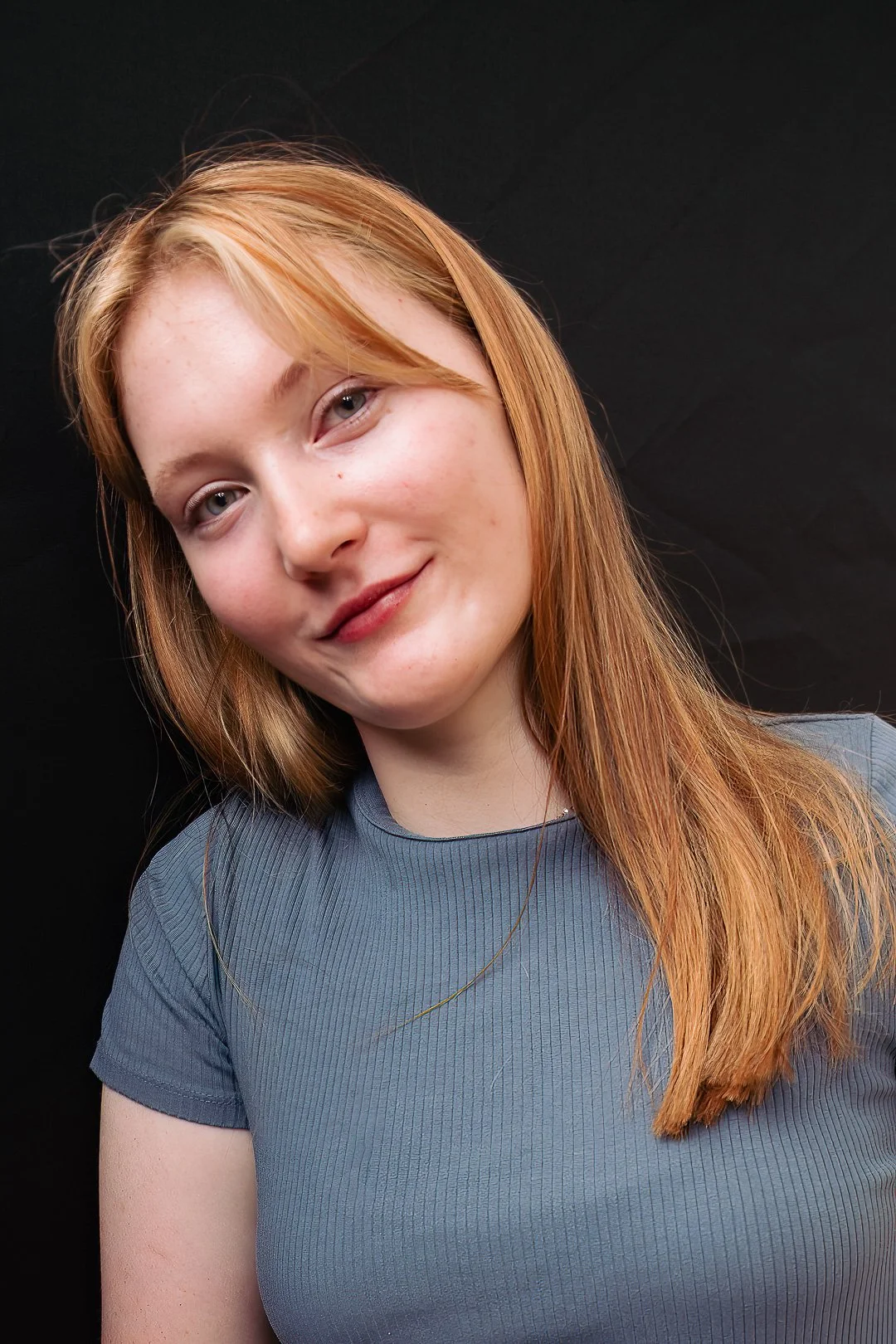 Close-up of a young woman with red hair wearing a blue top, smiling softly against a black background.