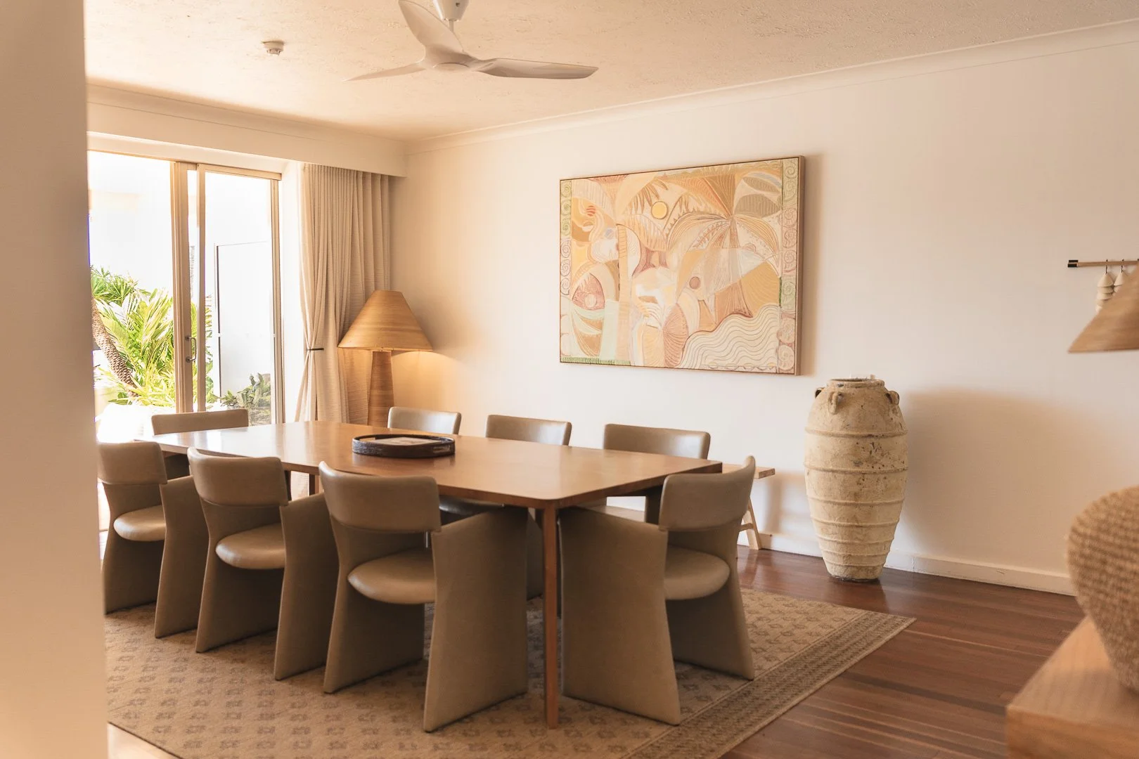 Dining room with wooden table and eight beige chairs, large ceramic vase, abstract painting on wall, sliding glass door with curtains, palm trees outside, ceiling fan, lamp with shade, and a patterned rug. Captured at Beach Club Resort, Hamilton Is