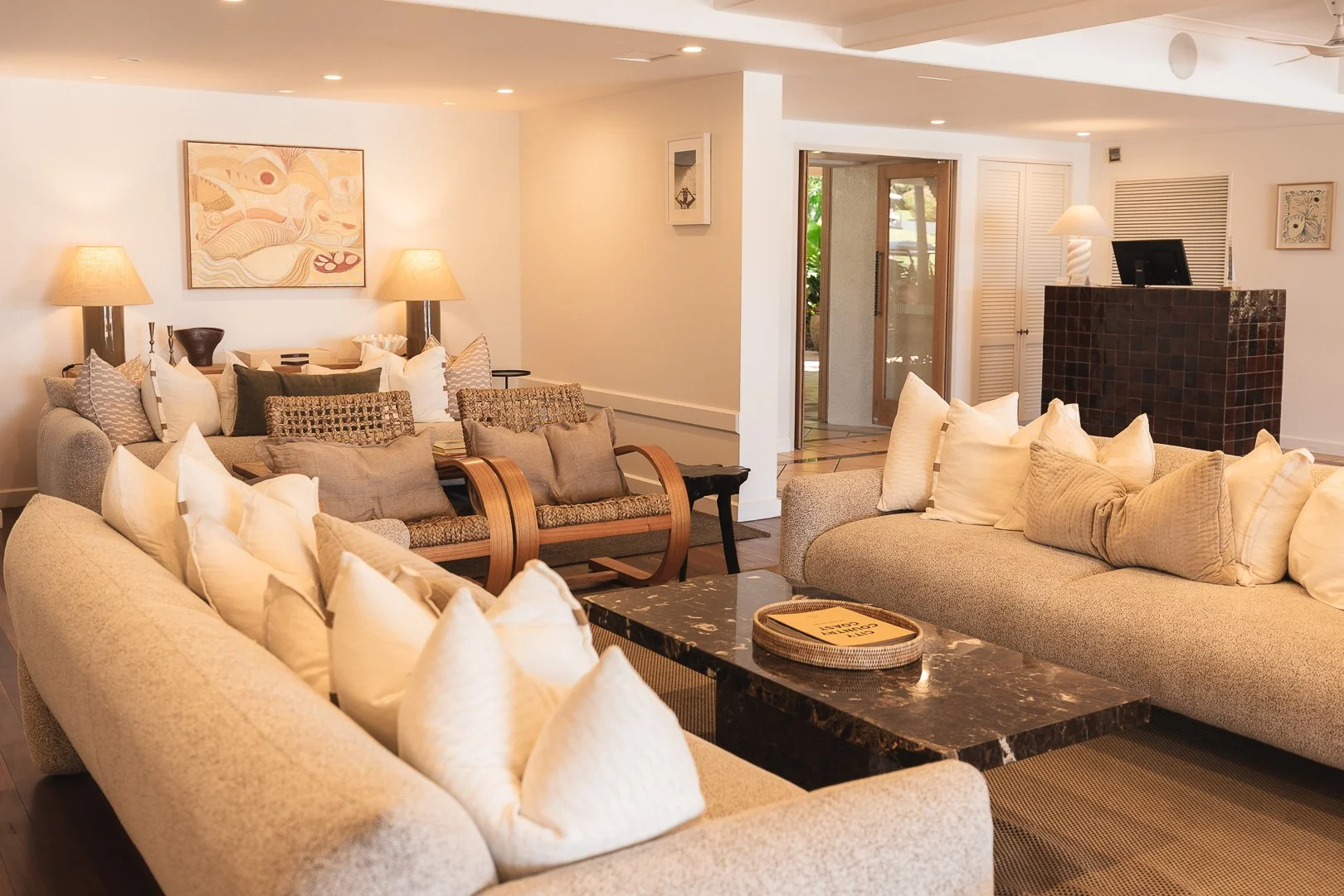 Interior of a cozy living room with beige couches, numerous pillows, a black marble coffee table, a wooden bench, and various lamps, with artwork on the walls and a door leading outside. Beach Club Whitsundays. Boutique accommodation photography