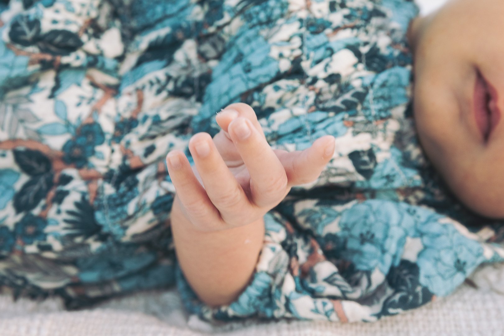 Close-up of a newborn baby’s hand resting on a floral blanket, with part of the baby's face visible.