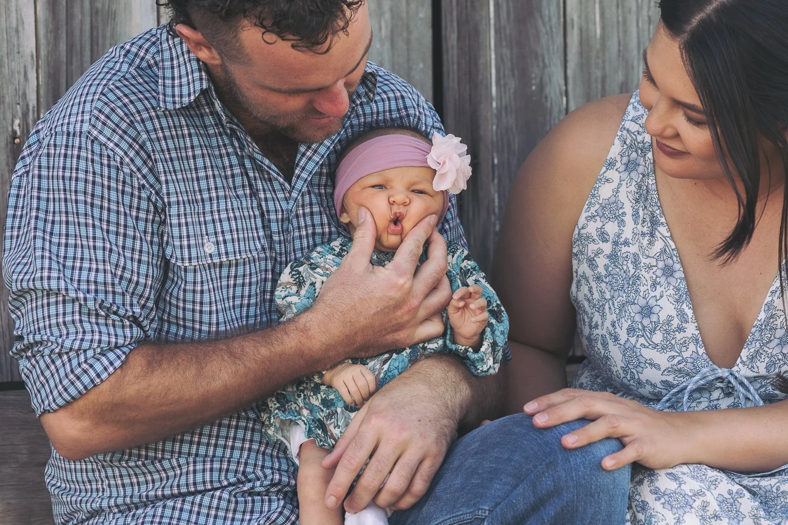 A man and woman sitting against a wooden fence holding a baby girl. The man is gently squeezing the baby's cheeks, making a funny face. The woman is smiling at them, wearing a sleeveless floral top. The baby girl is wearing a pink headband with a flo