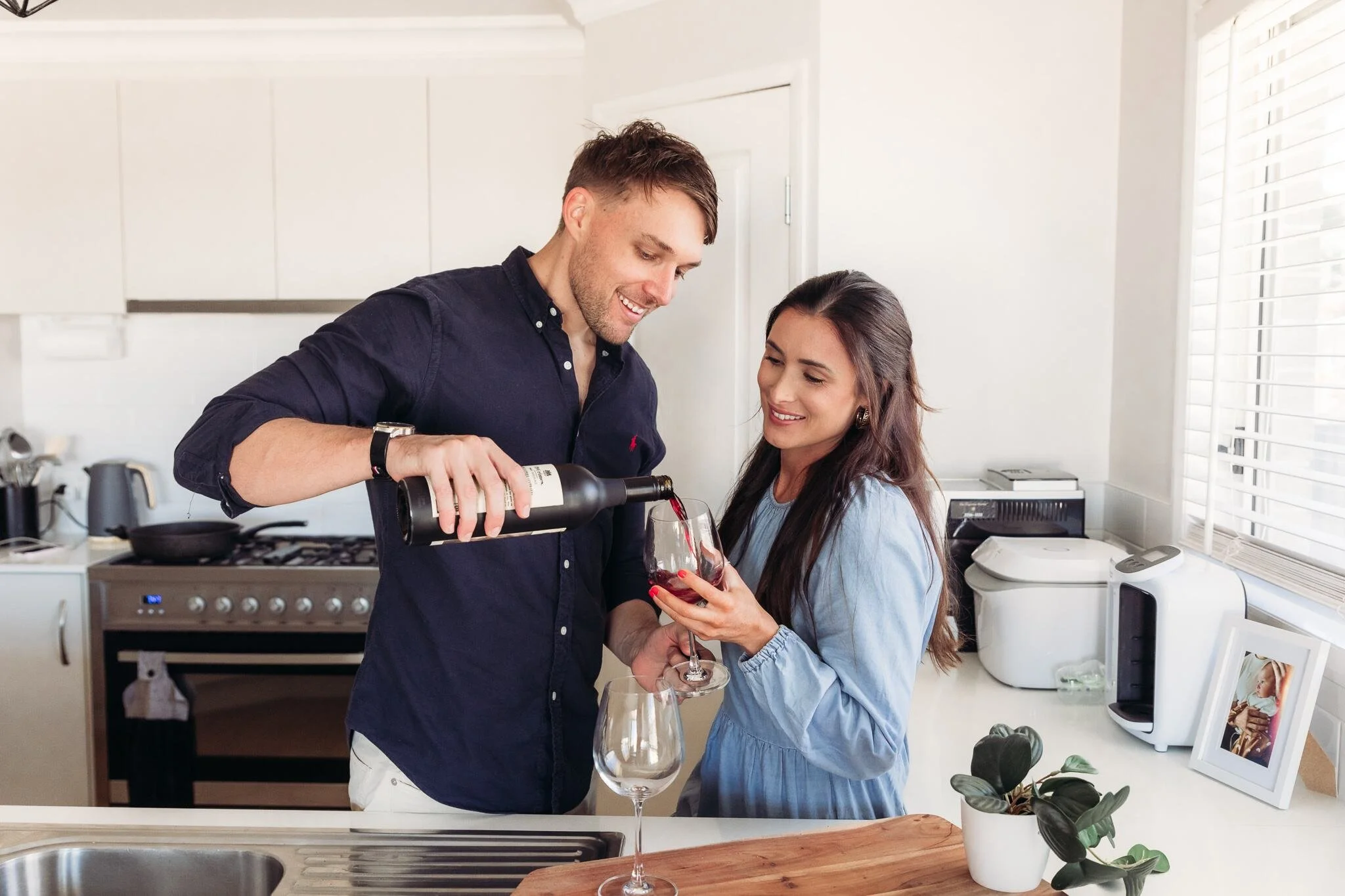 A man and woman in a kitchen, smiling as the man pours red wine into a wine glass held by the woman.