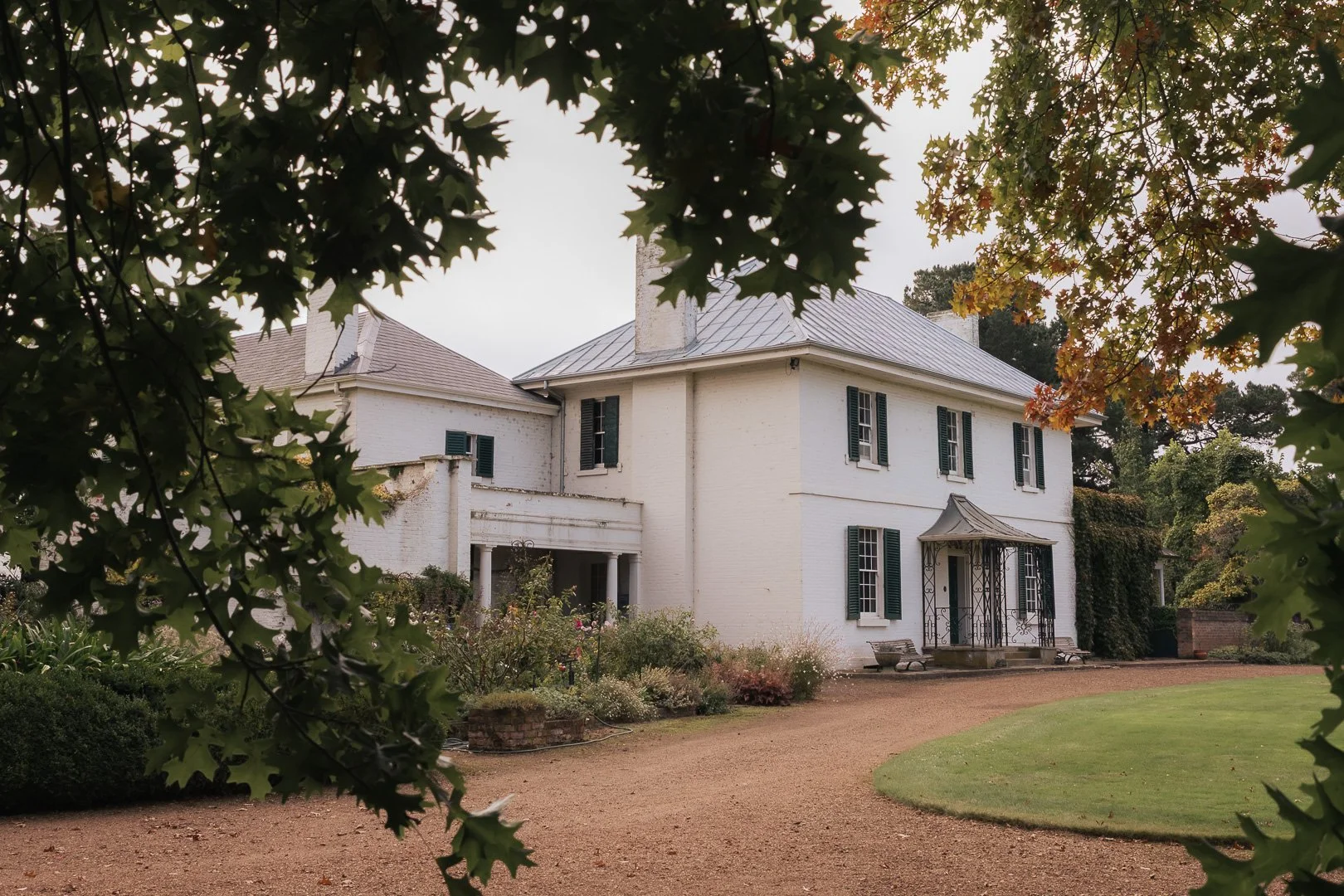 A large white house with green shutters, surrounded by trees and shrubbery, viewed through the branches of a tree with autumn leaves. House and Garden photography by Laurene Bardsley Photographer. Toowoomba Photographer