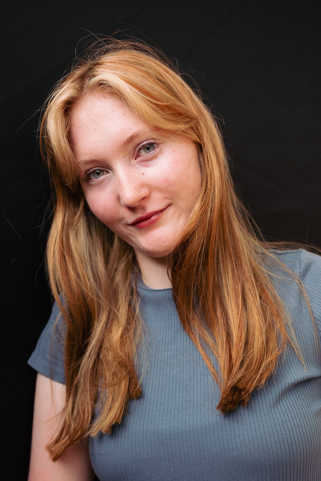 Portrait of a young woman with long red hair and blue eyes, wearing a gray shirt, smiling softly against a black background.