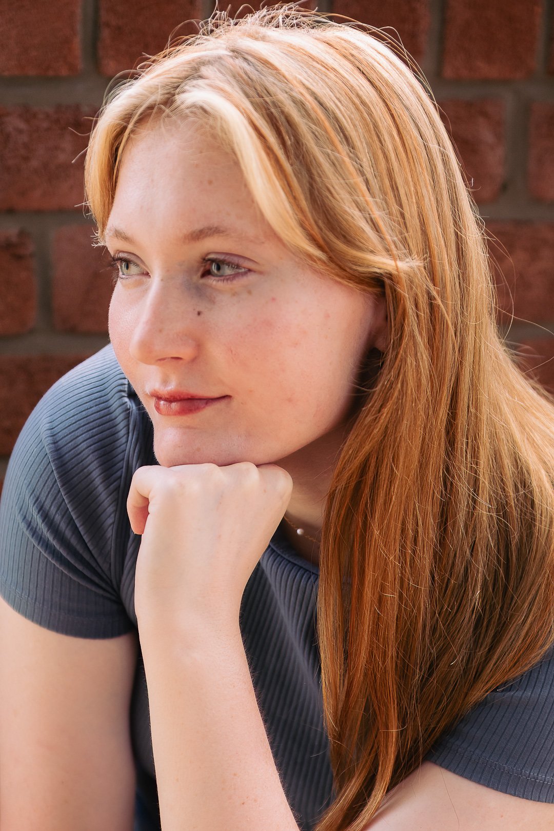 Close-up of a young woman with red hair and fair skin, resting her chin on her hand, looking thoughtfully to the side. She is wearing a dark gray shirt, and there is a brick wall in the background.