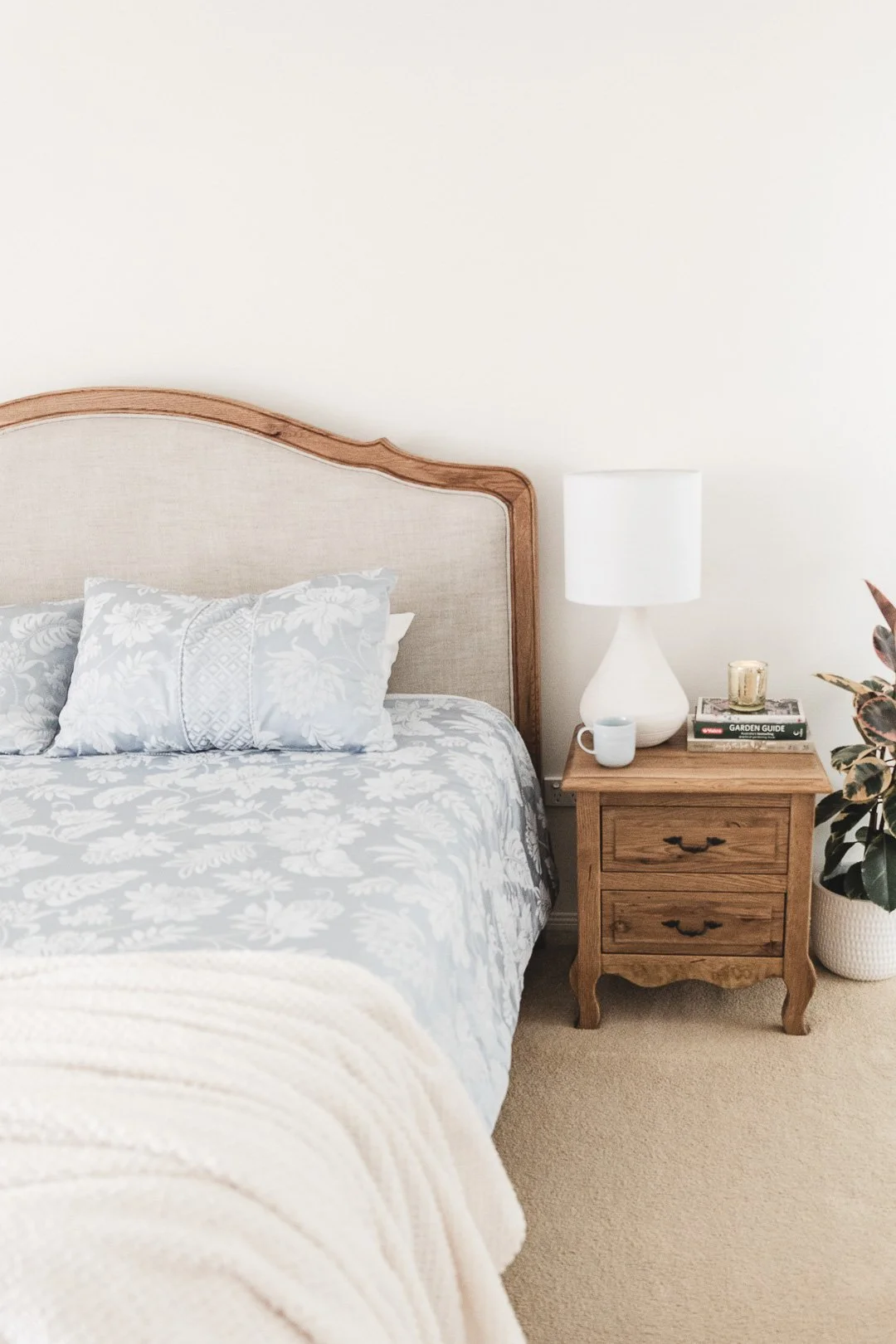 Bedroom with a beige headboard, gray and white floral bedding, wooden nightstand with a white lamp, books, a mug, and a candle, and a potted plant beside the nightstand.