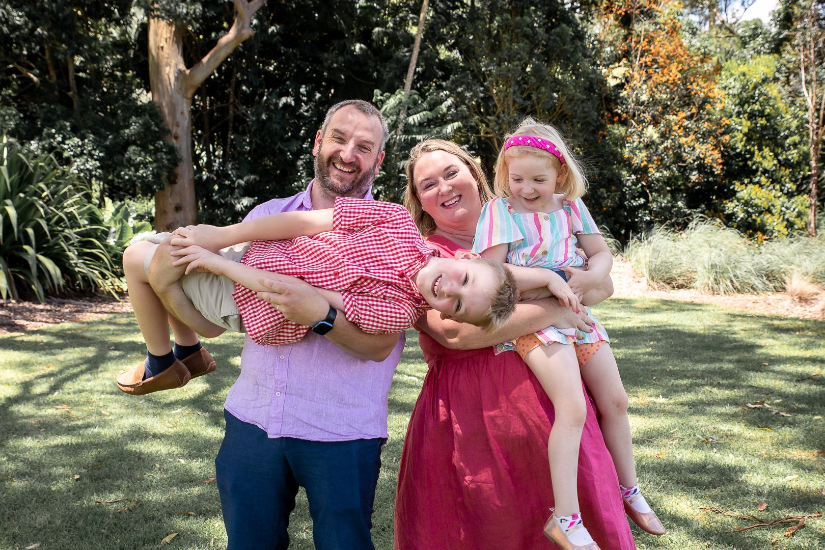 A family of four outdoors, with a man holding a boy and a woman holding a girl, over a grassy area with trees in the background.
