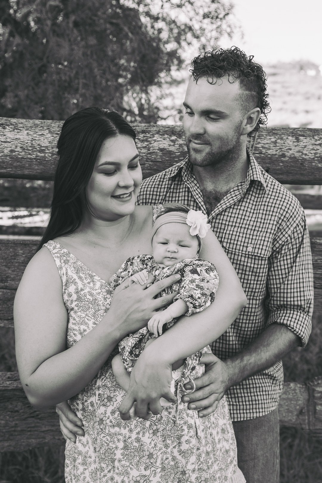 A family of three, mother, father, and baby, standing outdoors in front of a wooden fence. The mother is holding the baby, who has a bow headband, and both parents are looking at her affectionately.