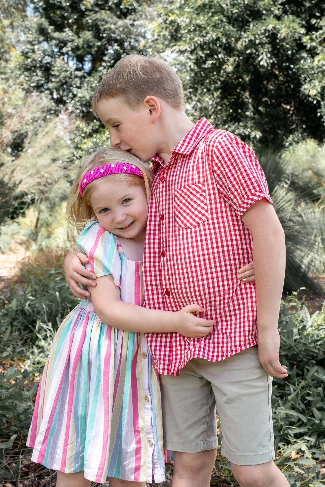 A boy and a girl hugging outdoors surrounded by greenery, with the boy kissing the girl's head.