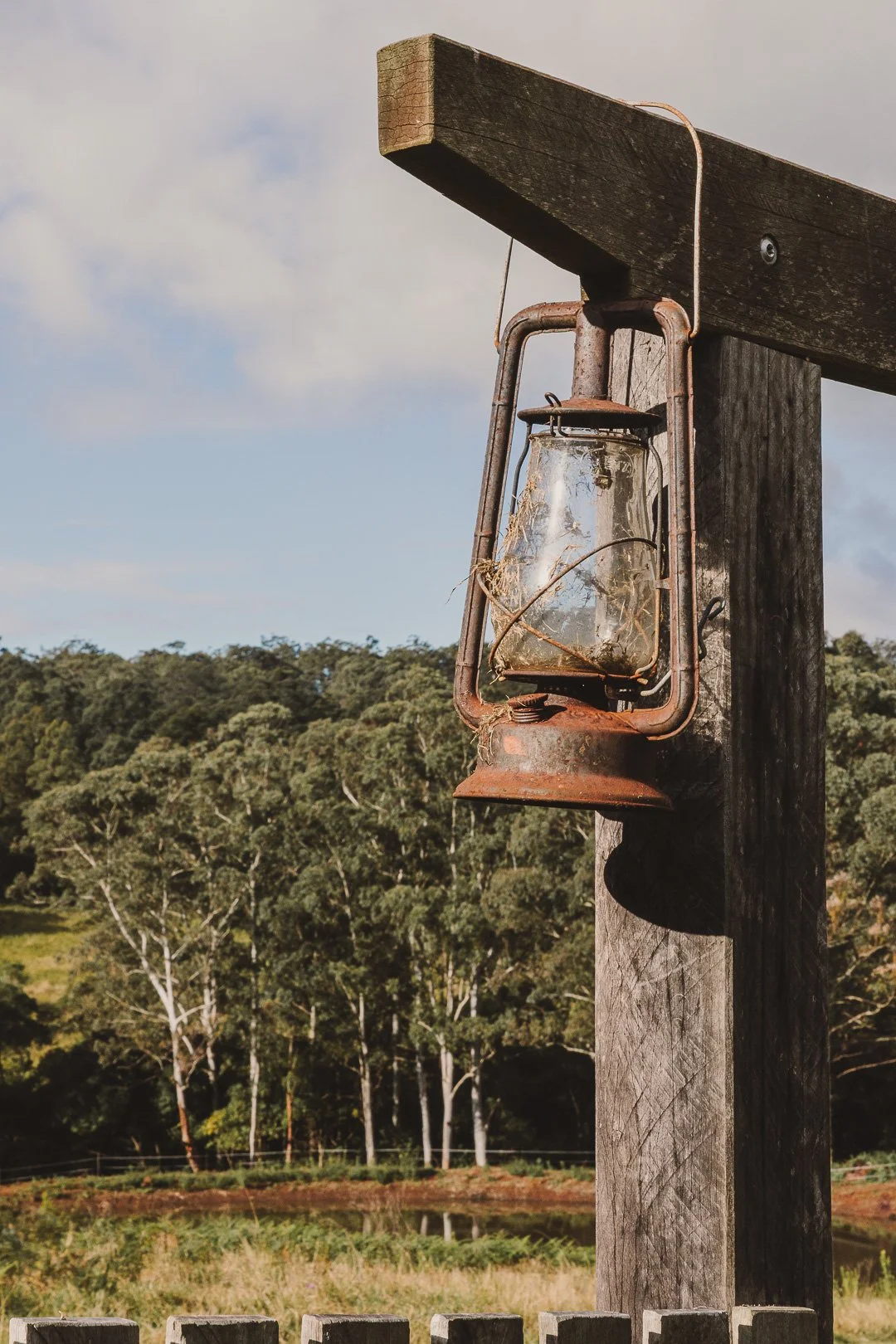 An old, rusty kerosene lantern hanging from a weathered wooden post outdoors with trees and a cloudy sky in the background.