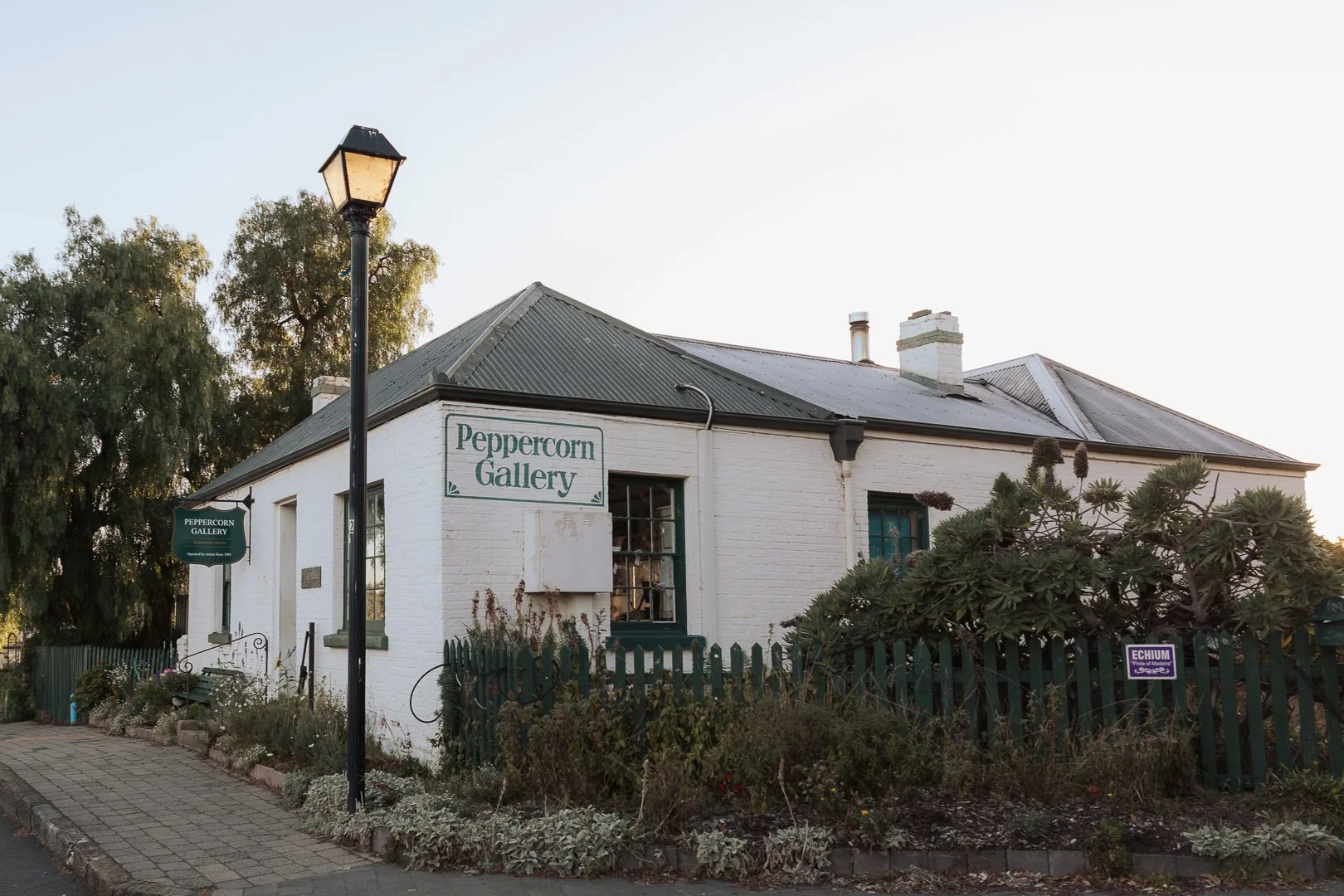 White brick building with green signs that read "Peppercorn Gallery," surrounded by plants and a green picket fence. A black street lamp stands in front of the building. The sky is clear and blue. Heritage photography by Laurene Bardsley Photography