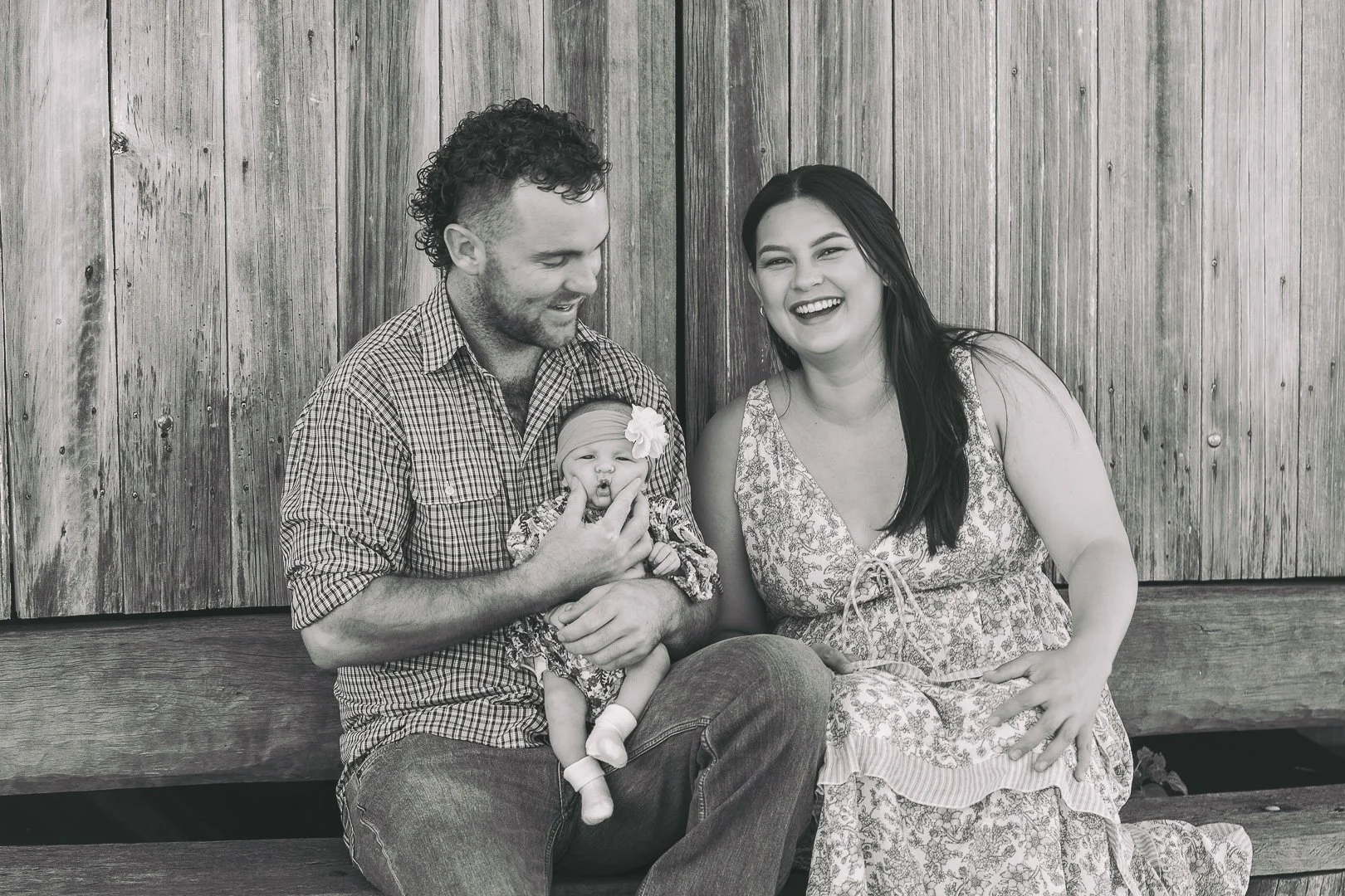A family of three, including a father, mother, and a baby girl, sitting on a bench in front of a wooden wall, smiling and looking at each other, with the father holding the baby.