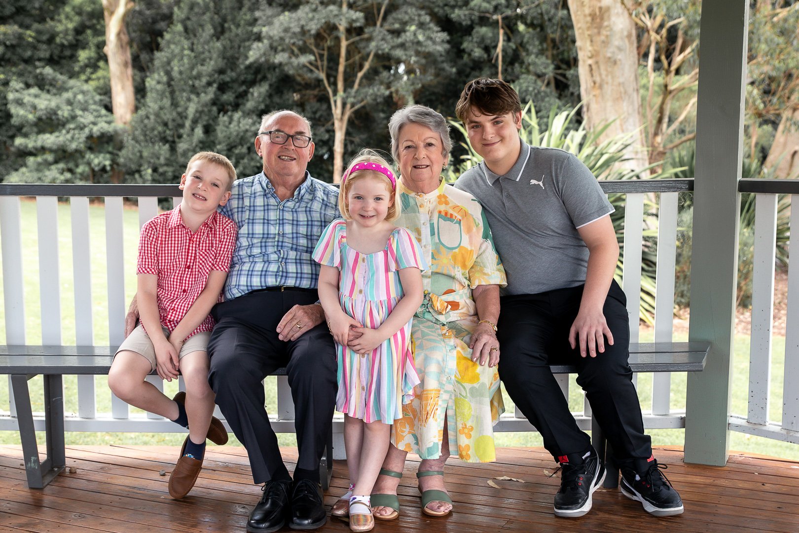Multi-generational family sitting on a porch bench outdoors, smiling, with trees in the background.