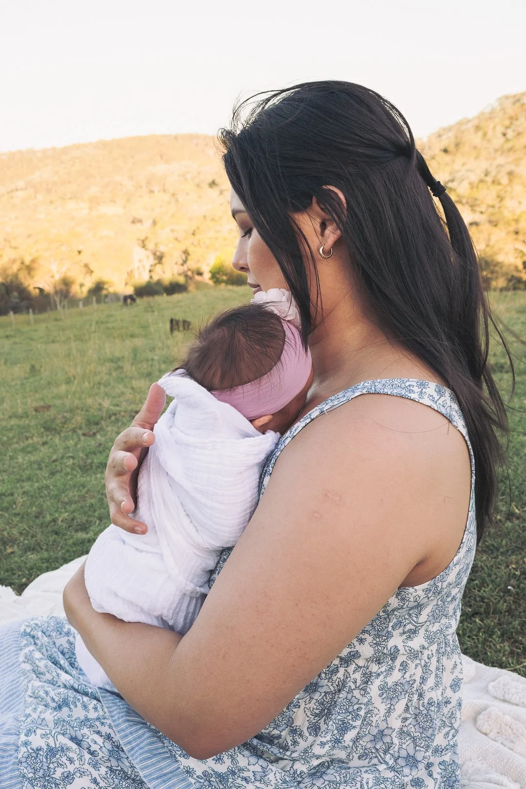 A woman holding a sleeping baby outdoors in a grassy field with hills in the background.