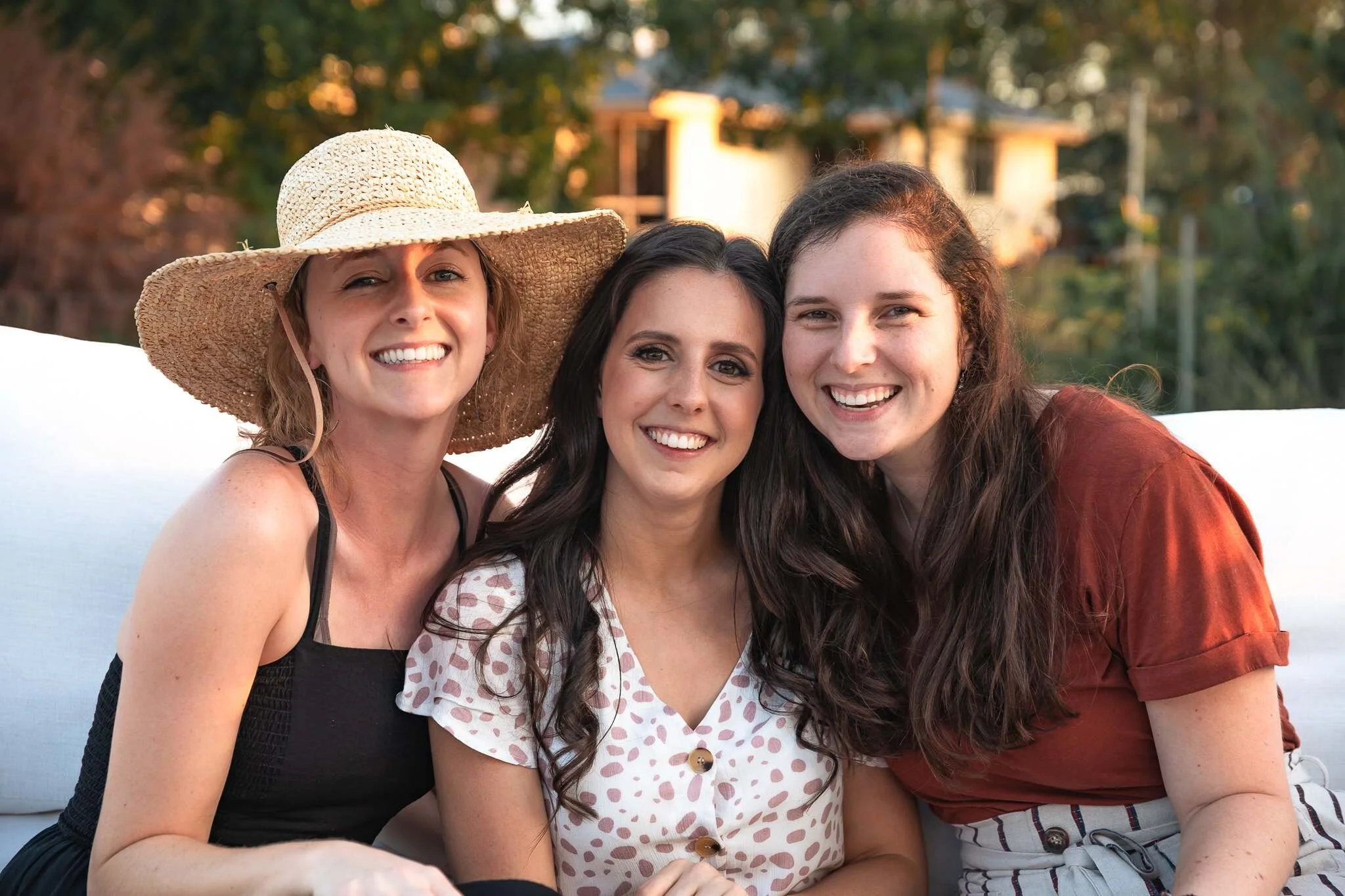 Three women smiling and sitting close together outdoors, with trees and a house in the background, during what appears to be late afternoon or early evening.