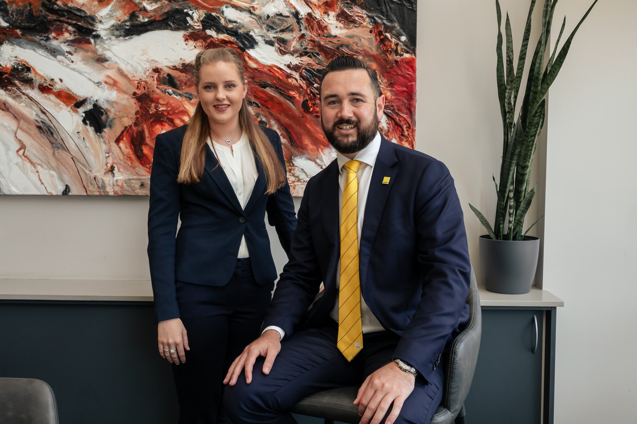 A woman and a man dressed in business attire in an office setting, with a large colorful abstract painting and a tall potted plant in the background.