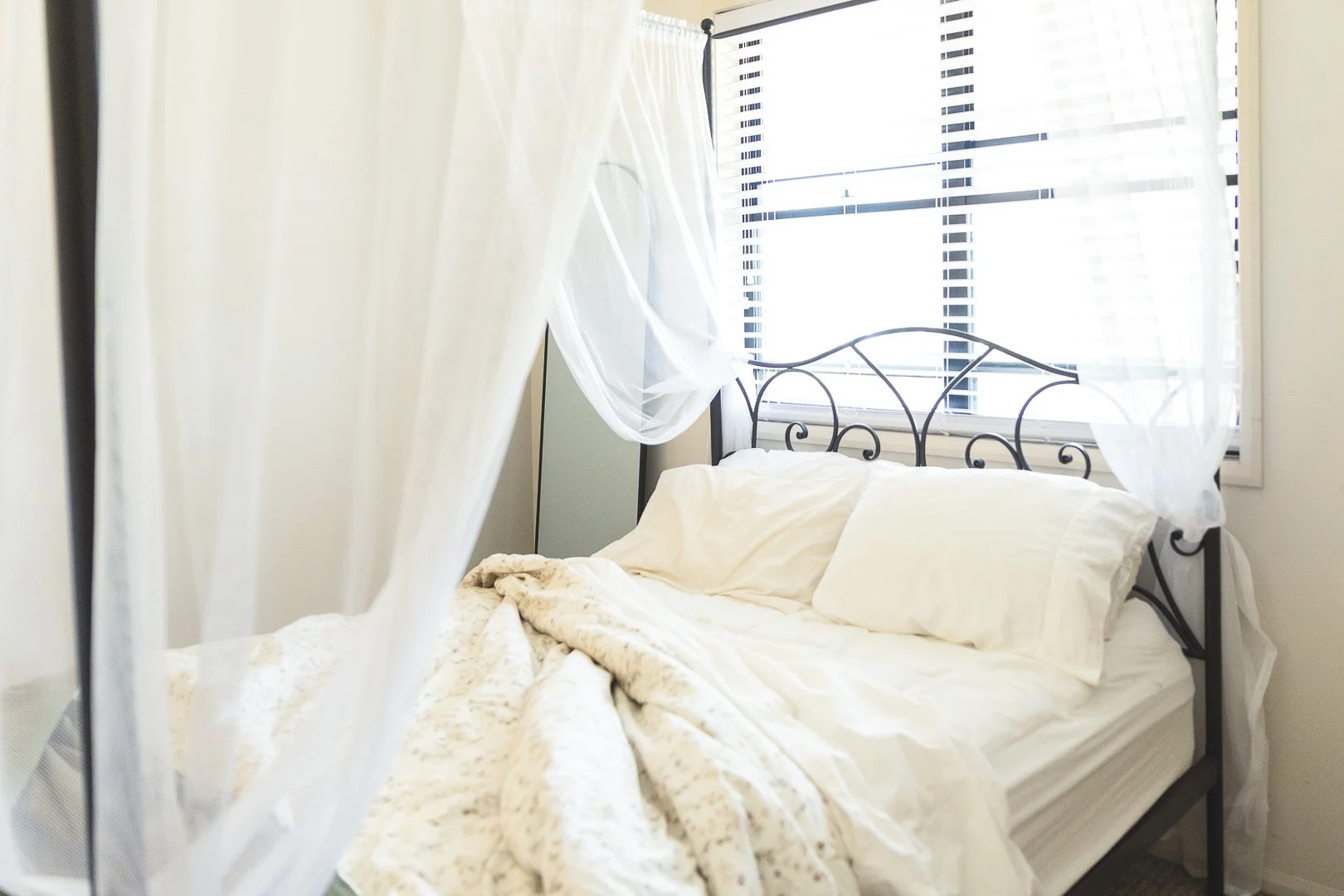 A bed with white bedding and pillows in a room with large windows with white blinds and sheer curtains, and a black wrought iron headboard.