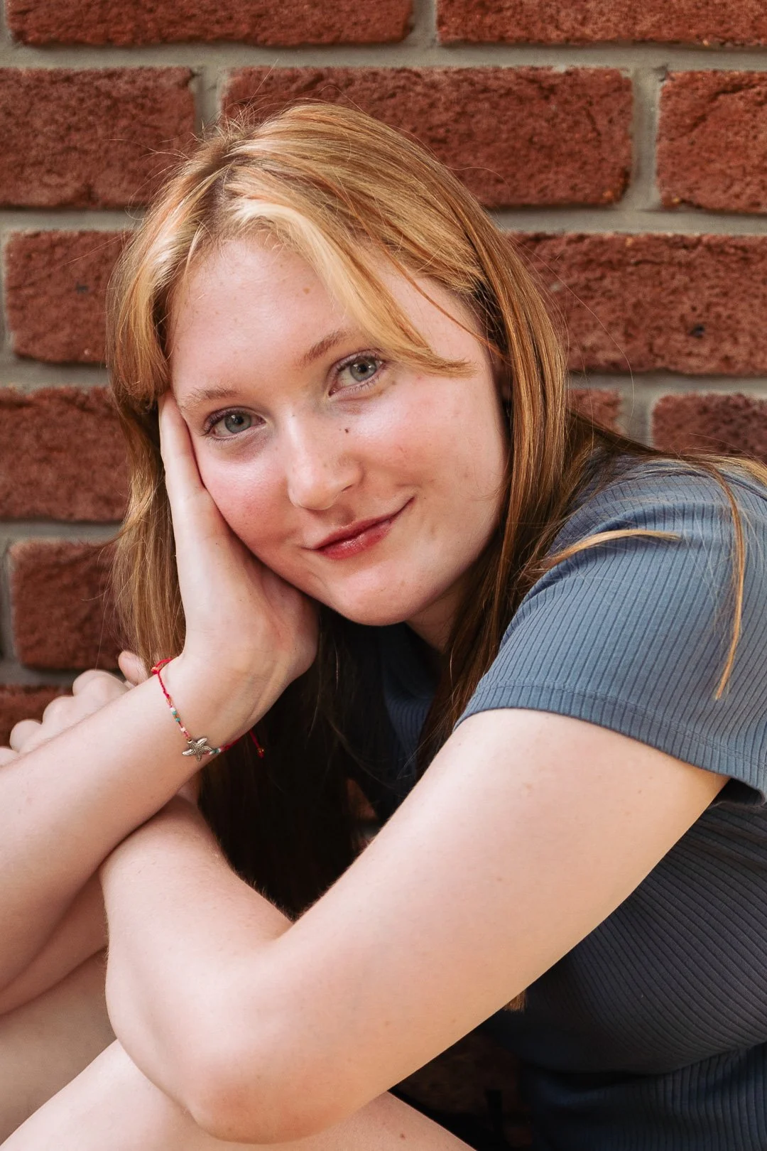 A young woman with long red hair, blue eyes, and freckles, resting her head on her hand and smiling, sitting against a brick wall.