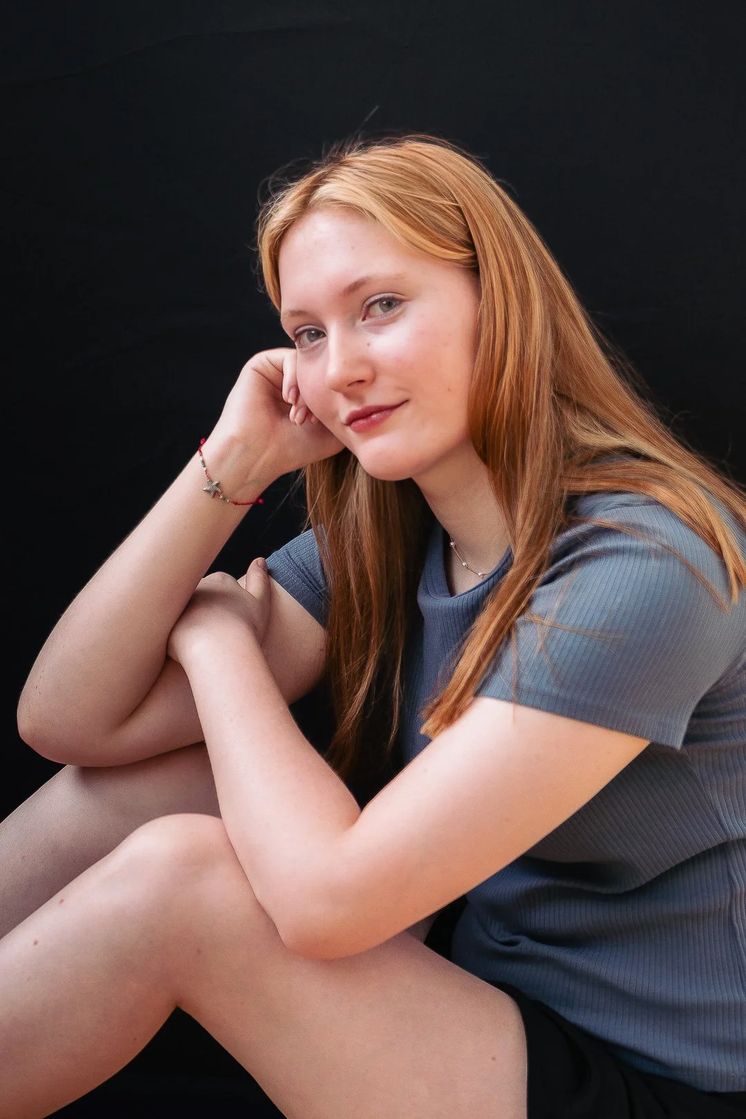 A young woman with long red hair sitting against a black background, resting her head on her hand and smiling softly at the camera.