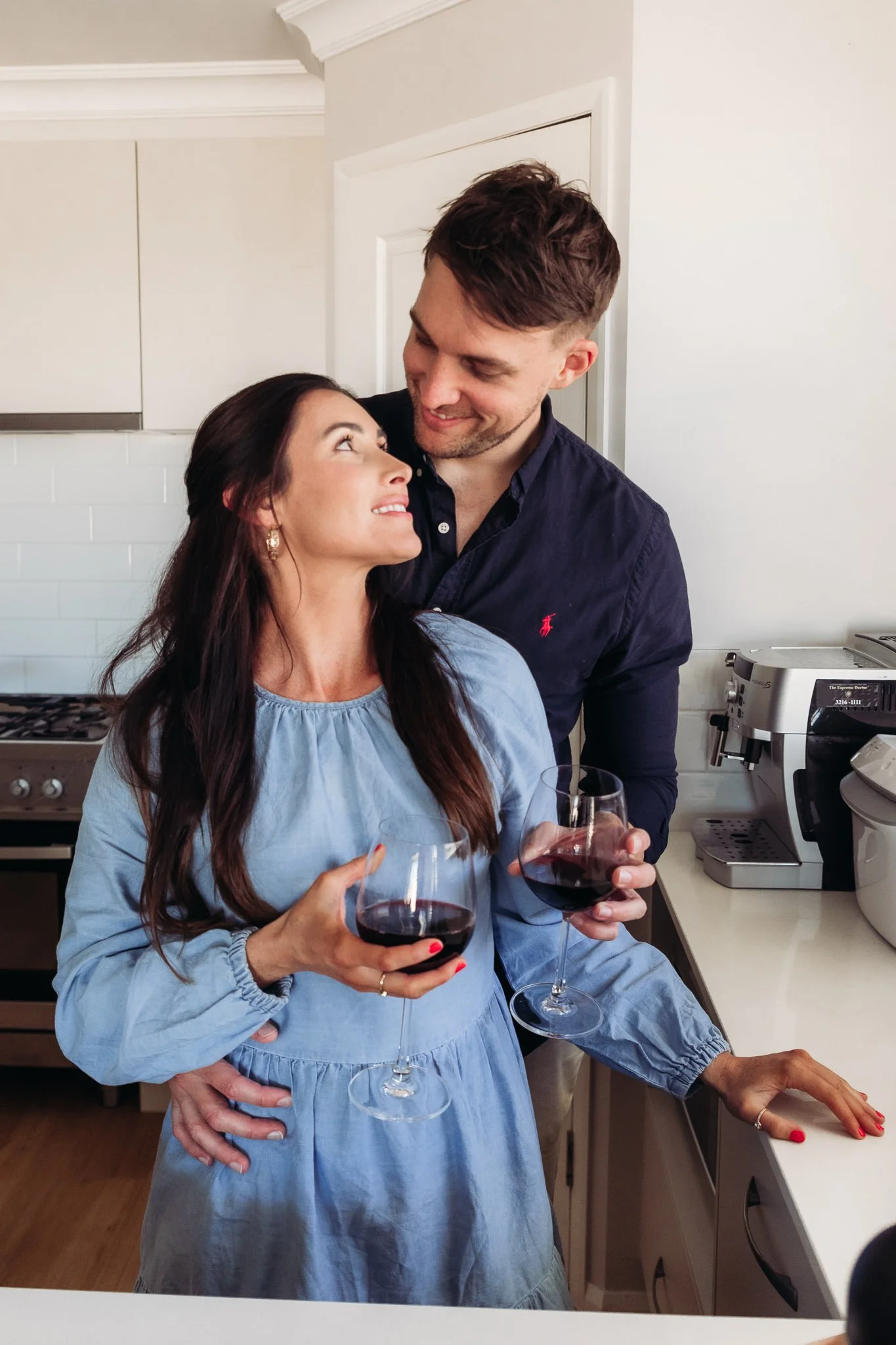 A man and woman in a kitchen, smiling and looking at each other, holding glasses of red wine.