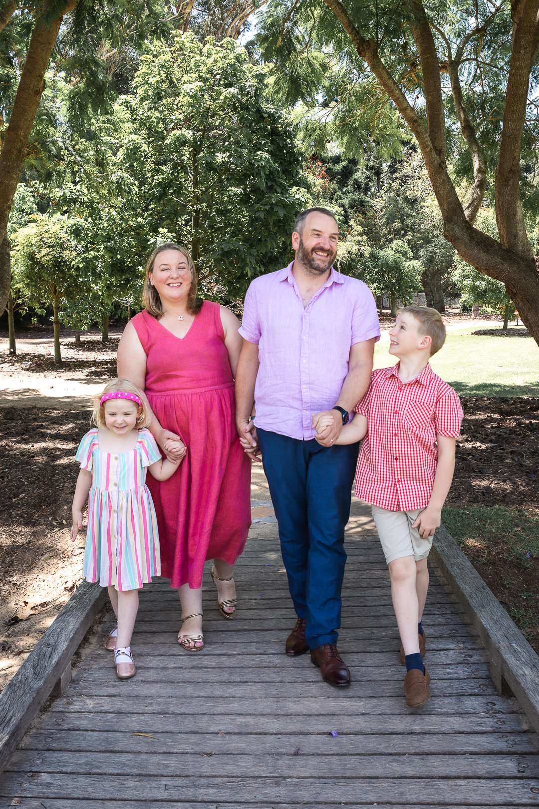 Family walking on a wooden bridge in a park with trees, holding hands and smiling.