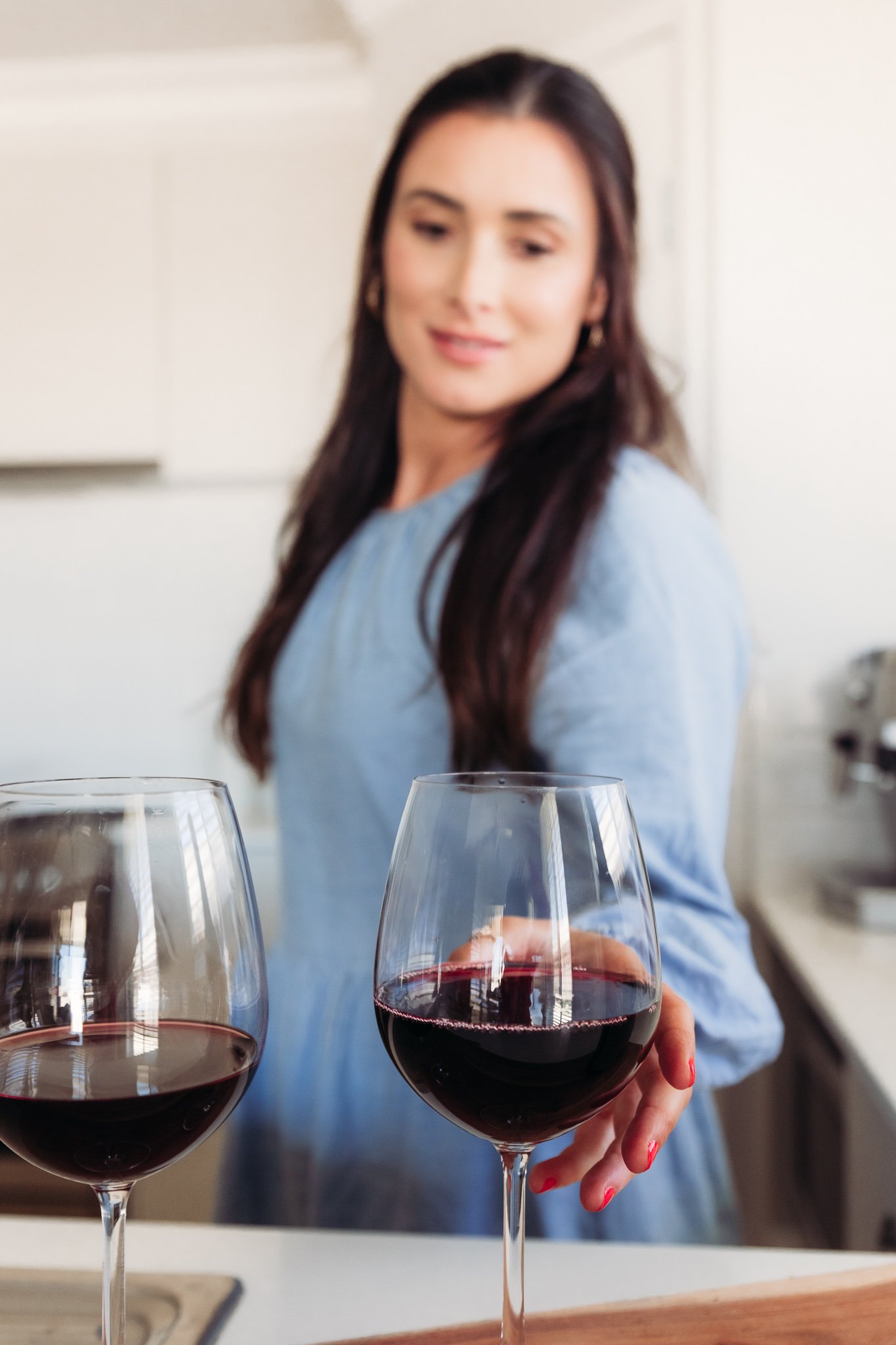 A woman with long dark hair holding a glass of red wine in a kitchen, with another glass of red wine on the counter in front of her.