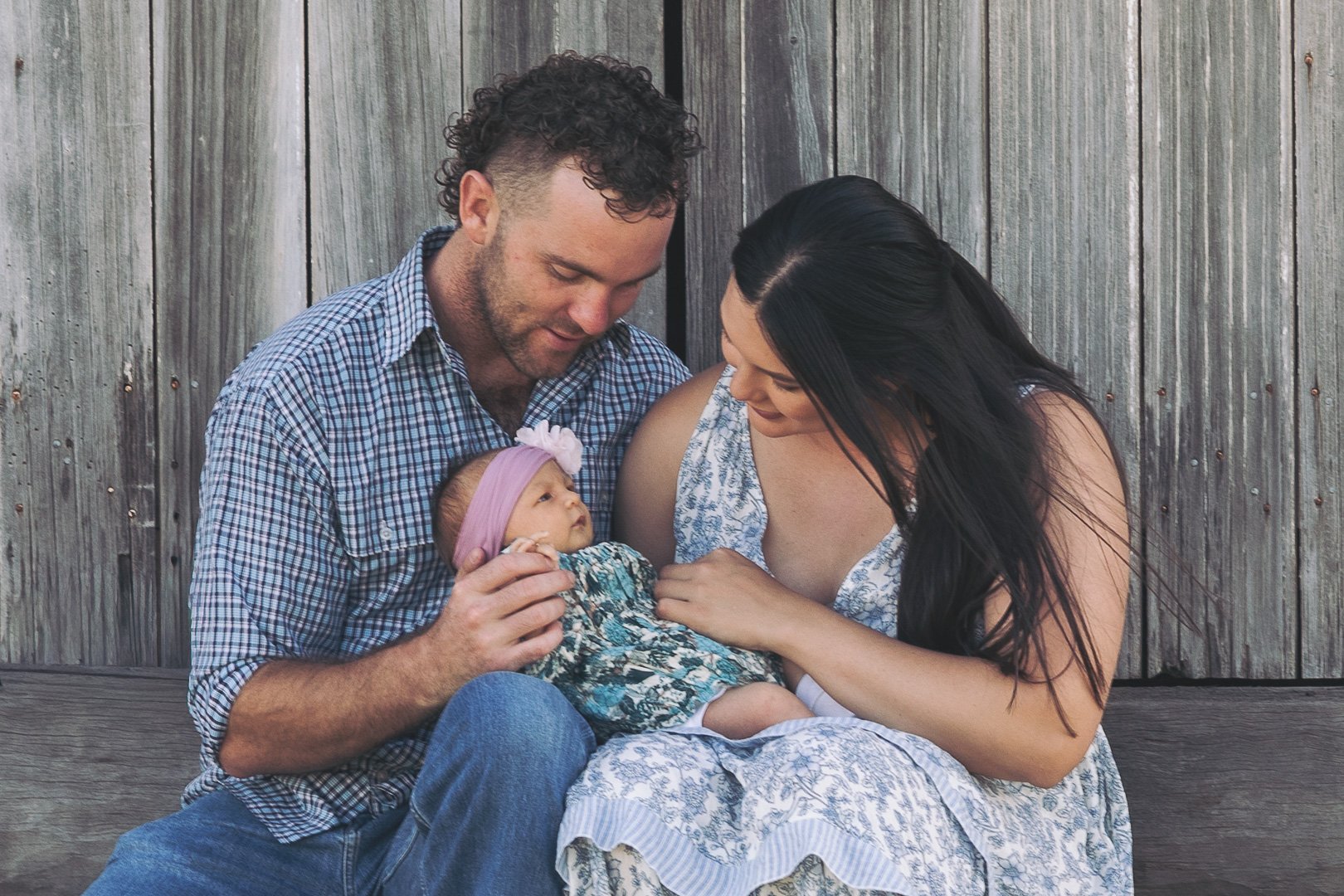 A man and woman sit close together holding a baby girl, looking lovingly at her, against a wooden fence background.