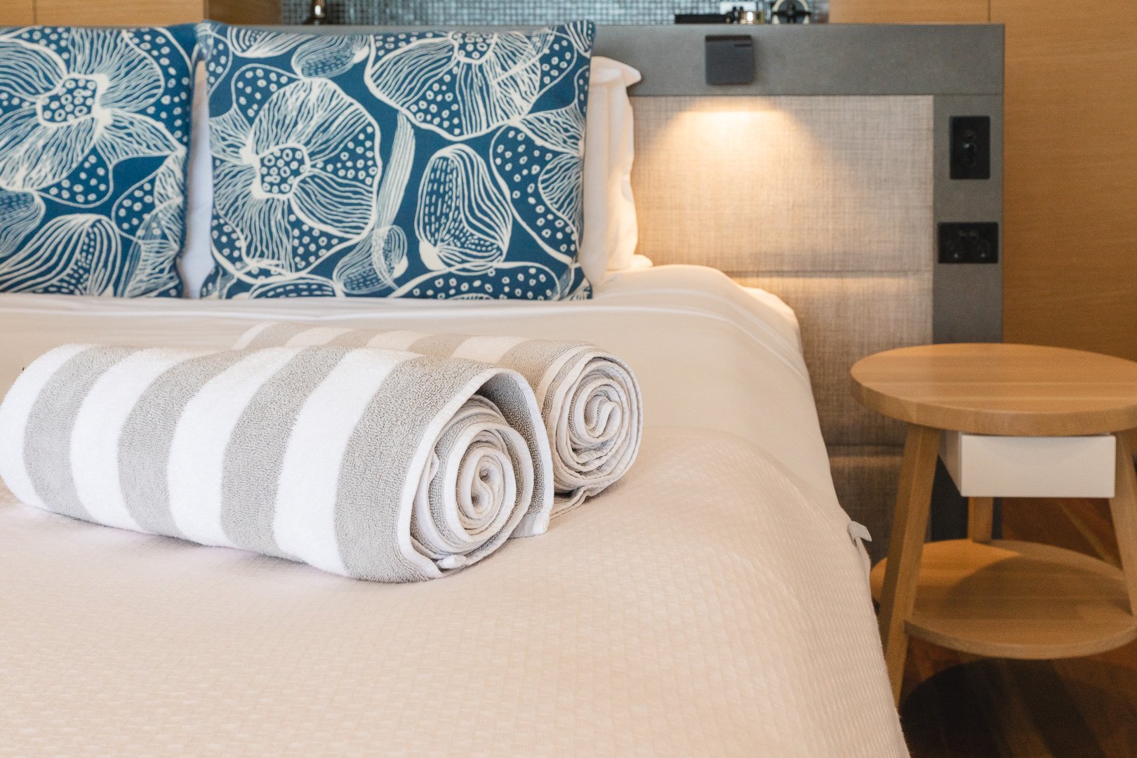 A neatly made hotel bed with two rolled white and gray striped towels, two decorative blue pillows with white floral patterns, a beige headboard with electrical outlets, and a round wooden side table. Beach Club Hotel, Hamilton Island, Queensland. 