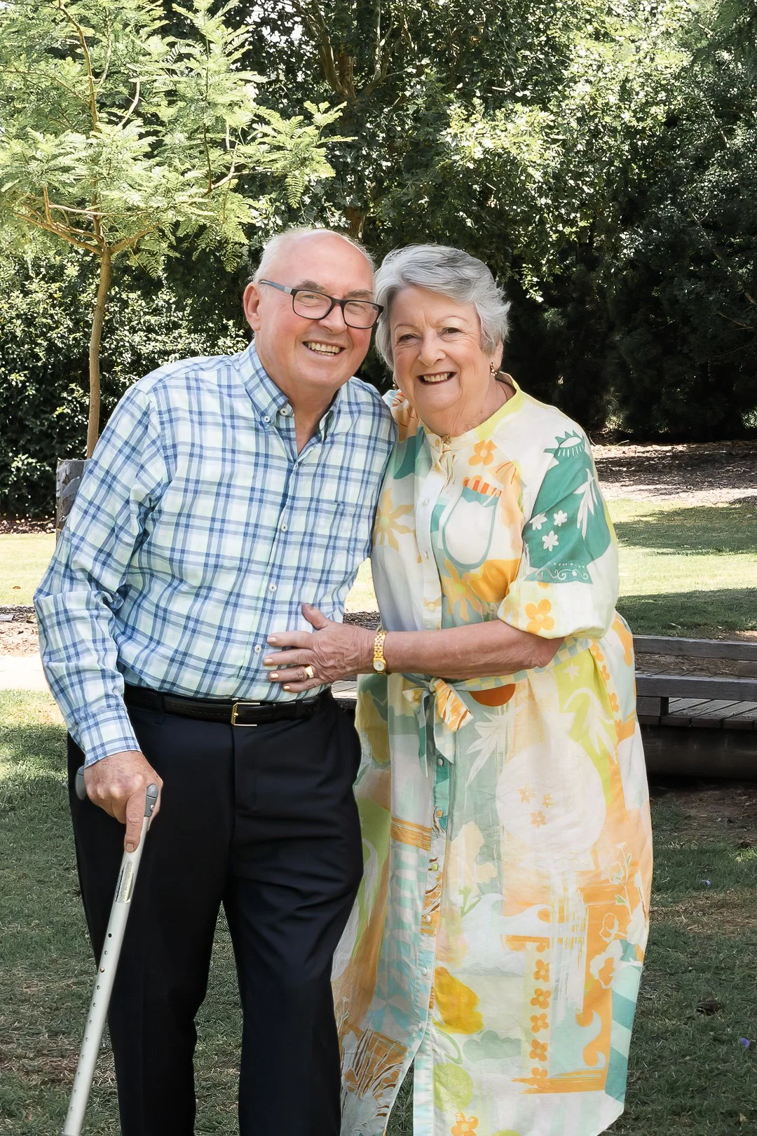 An elderly man and woman smiling and standing together outdoors, with the man holding a cane, surrounded by trees and grass.