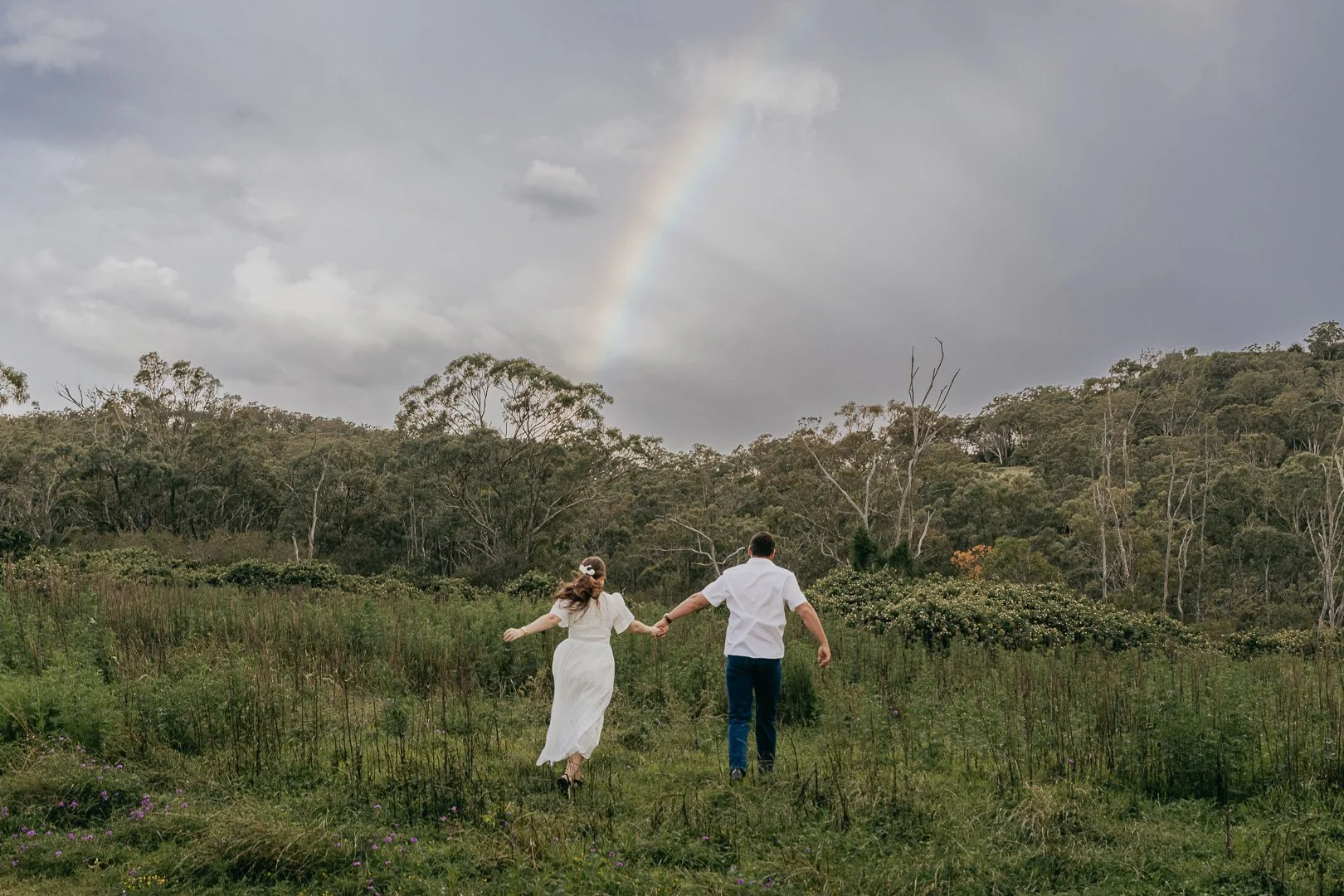 A couple holding hands, walking through a grassy field with trees and bushes, a rainbow and cloudy sky in the background.