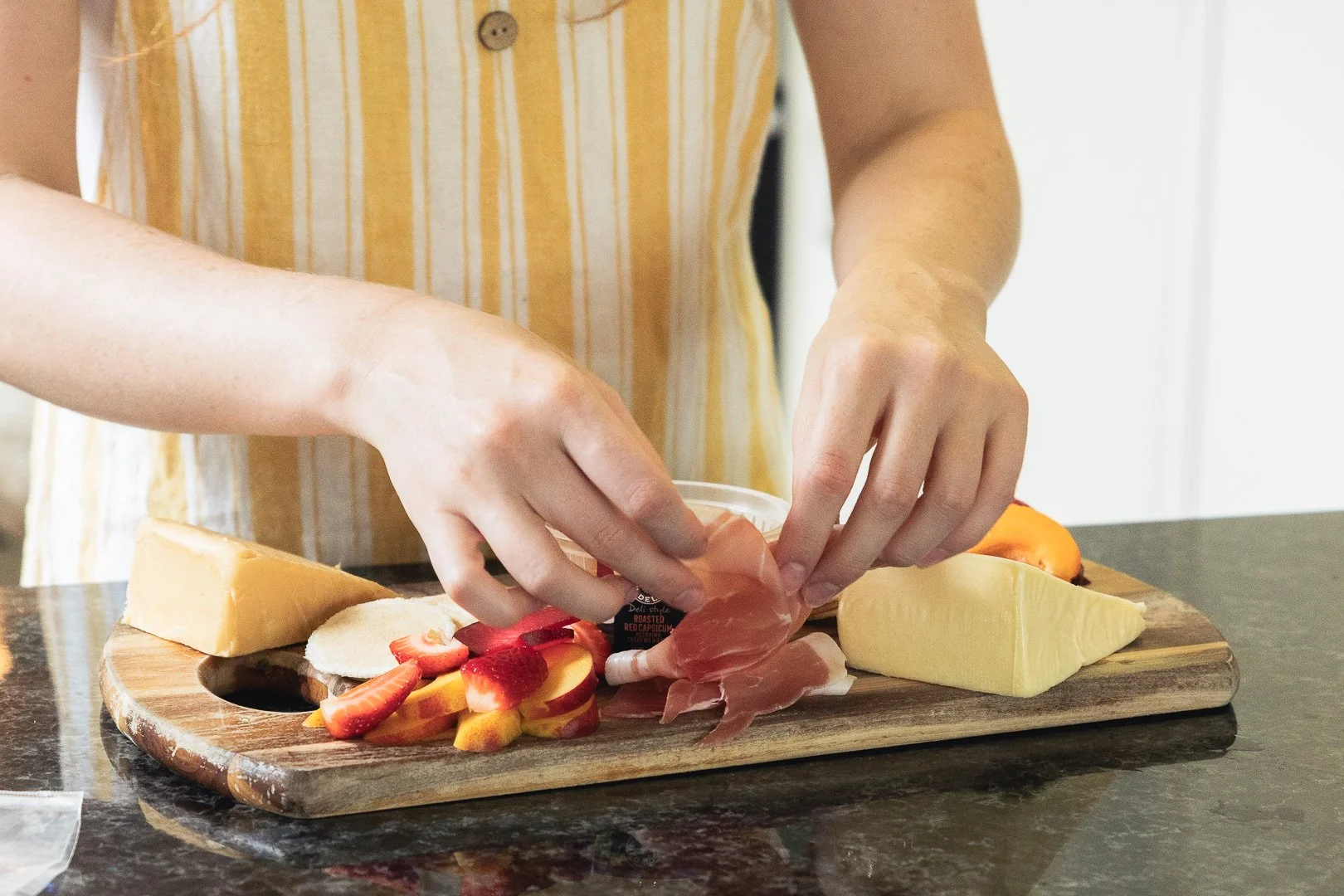 Person preparing a cheese and fruit platter with sliced strawberries, peaches, prosciutto, and cheese on a wooden cutting board.