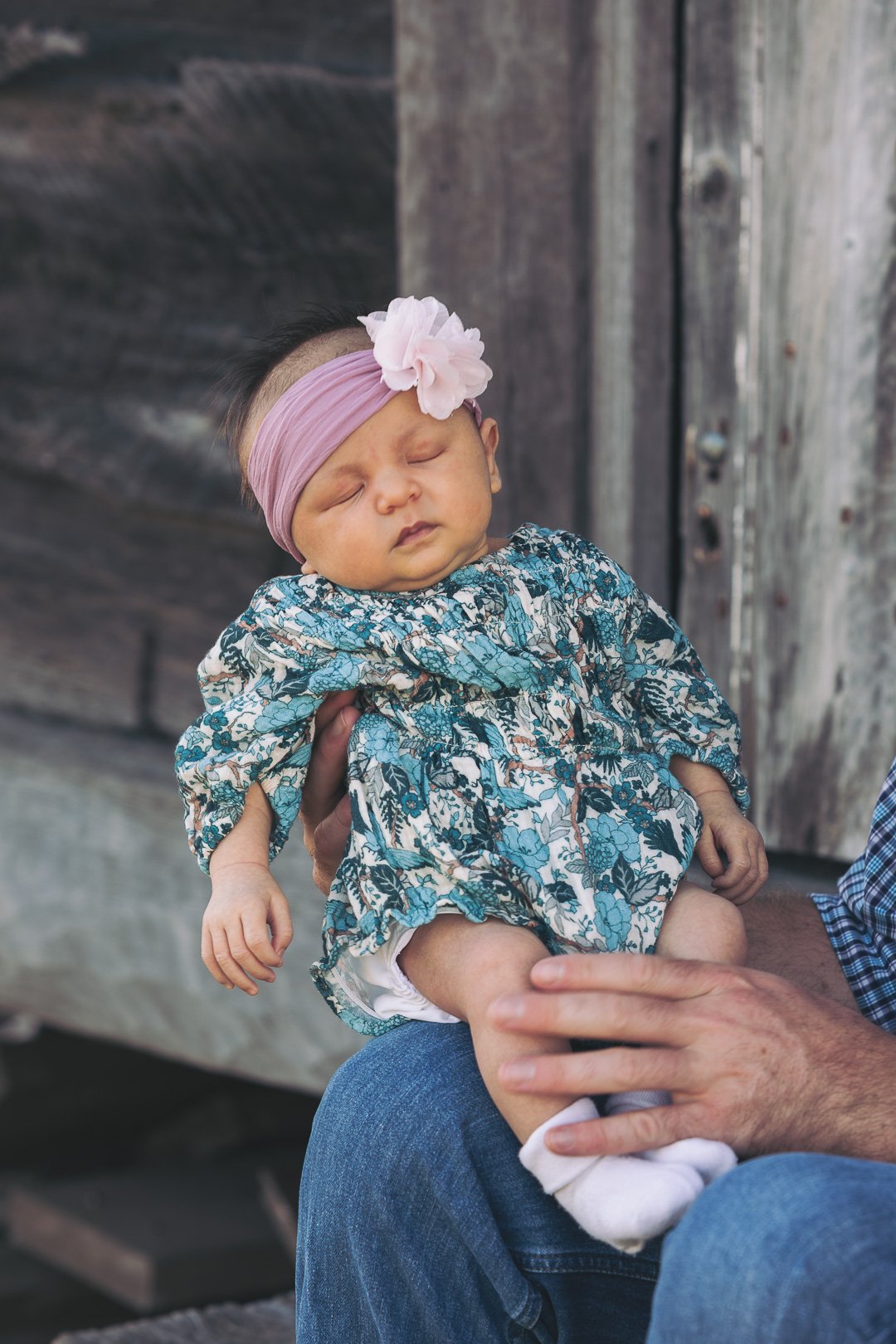 A sleeping baby girl wearing a pink headband with a large flower, a floral dress, and white socks, sitting on an adult's lap outdoors near wooden structures.