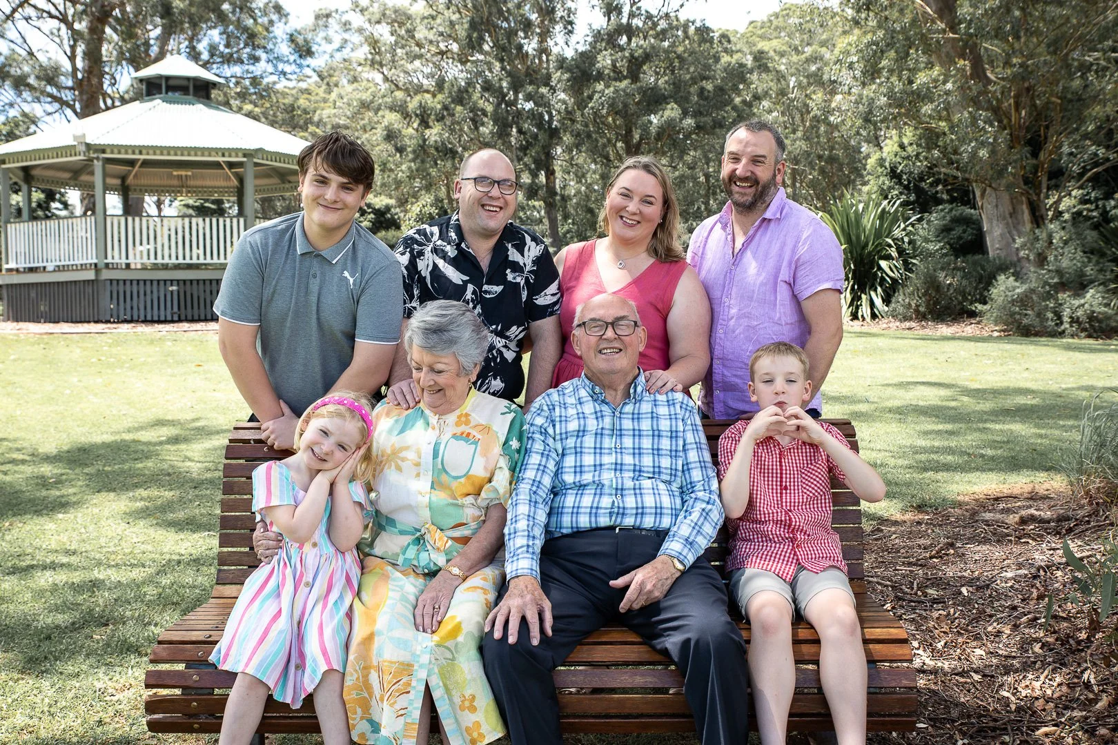 Family group photo outdoors in a park, with three generations, sitting and standing around a wooden bench, smiling, surrounded by trees and grass.