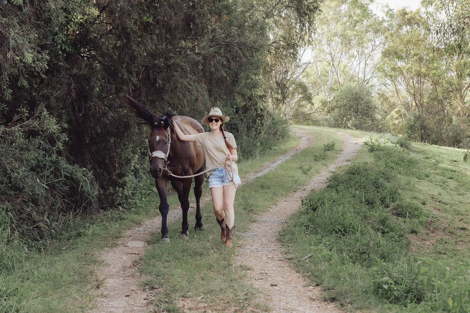 A woman in a beige t-shirt, denim shorts, and cowboy boots walking alongside a brown horse on a dirt trail in a lush green forest.