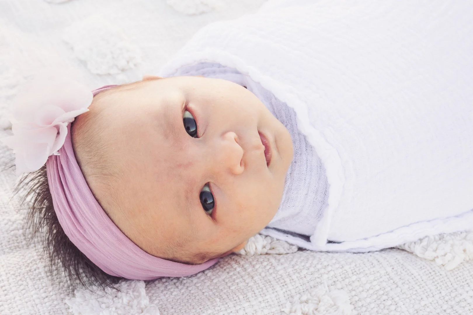 Close-up of a newborn baby girl with a pink headband and bow, lying on a white textured blanket, wearing a white outfit, with clear eyes looking at the camera.