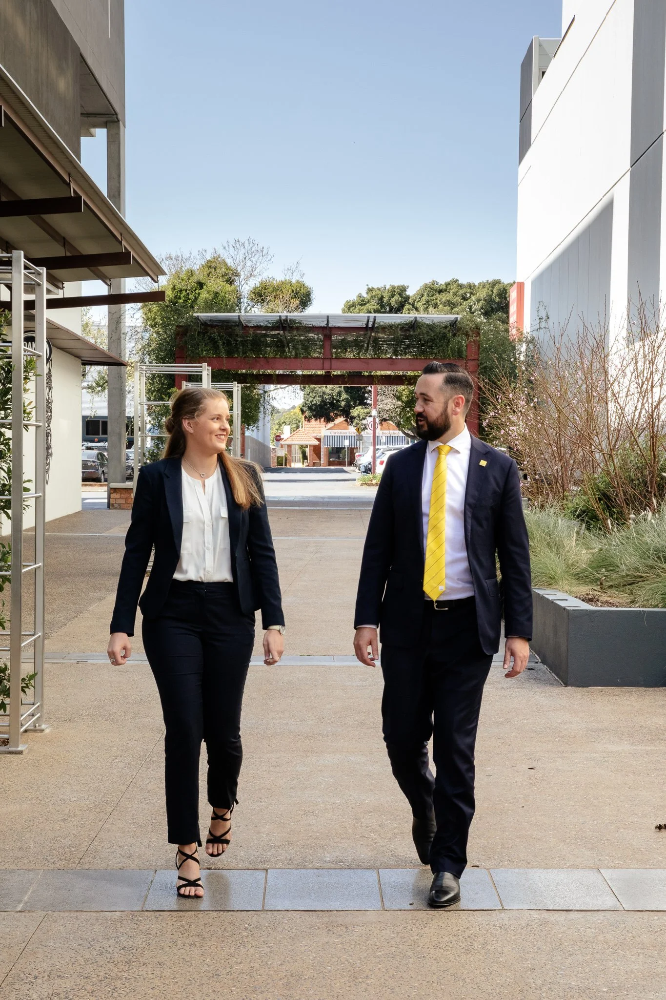A man and woman walking and talking outdoors, both dressed in business attire, with the woman smiling and the man looking at her. An example of a branding photography session.